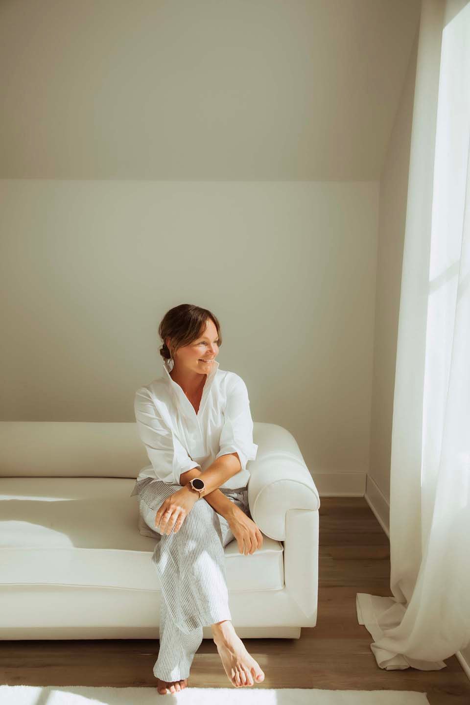 A smiling woman in a white robe sits barefoot on a cream sofa in a bright, sunlit room near a sheer curtain.