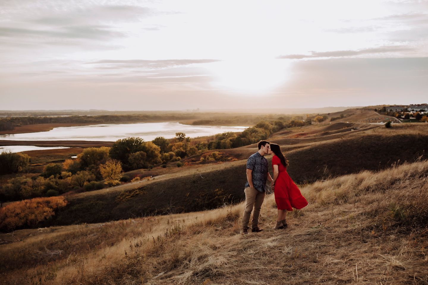 Engaged couple stand together on a minnesotan hillside overlooking a scenic lake at sunset.