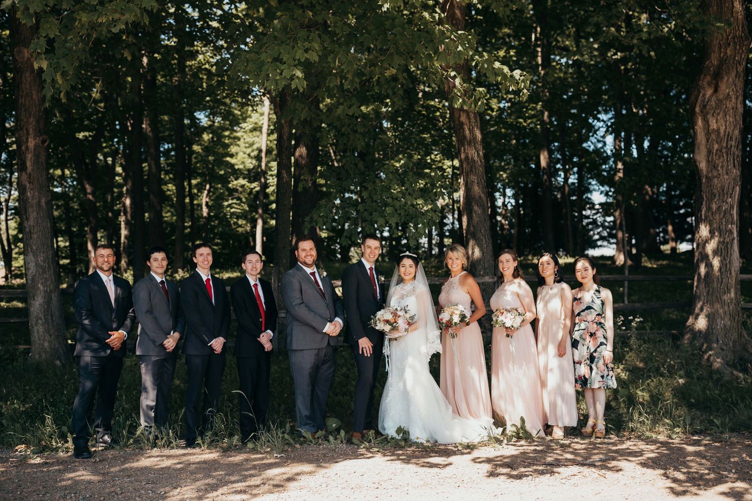 Wedding party stands in line against a wooded backdrop wearing navy suits and blush dresses.