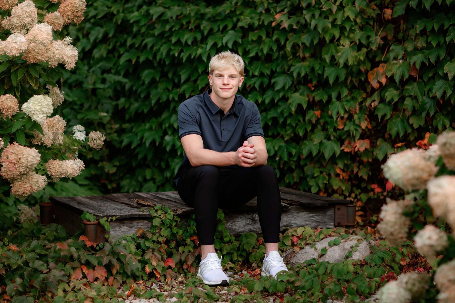 Casual portrait seated outdoors with hydrangea bushes and green foliage backdrop.