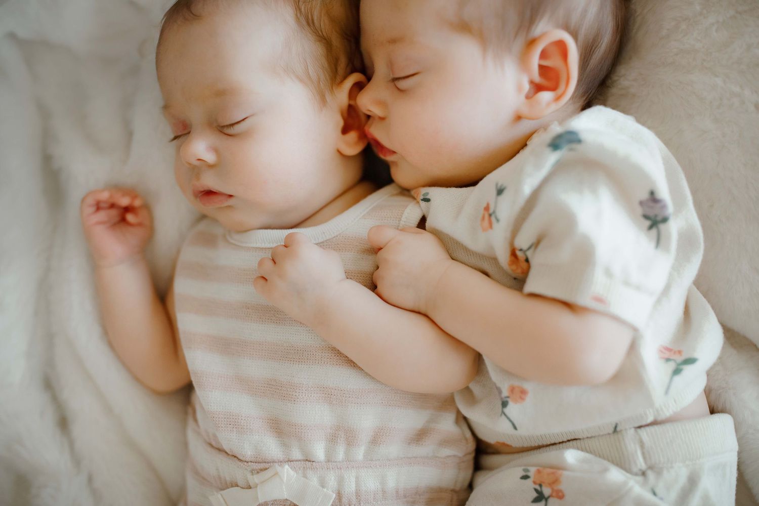 Two sleeping babies in light colored outfits lying close together on white bedding.