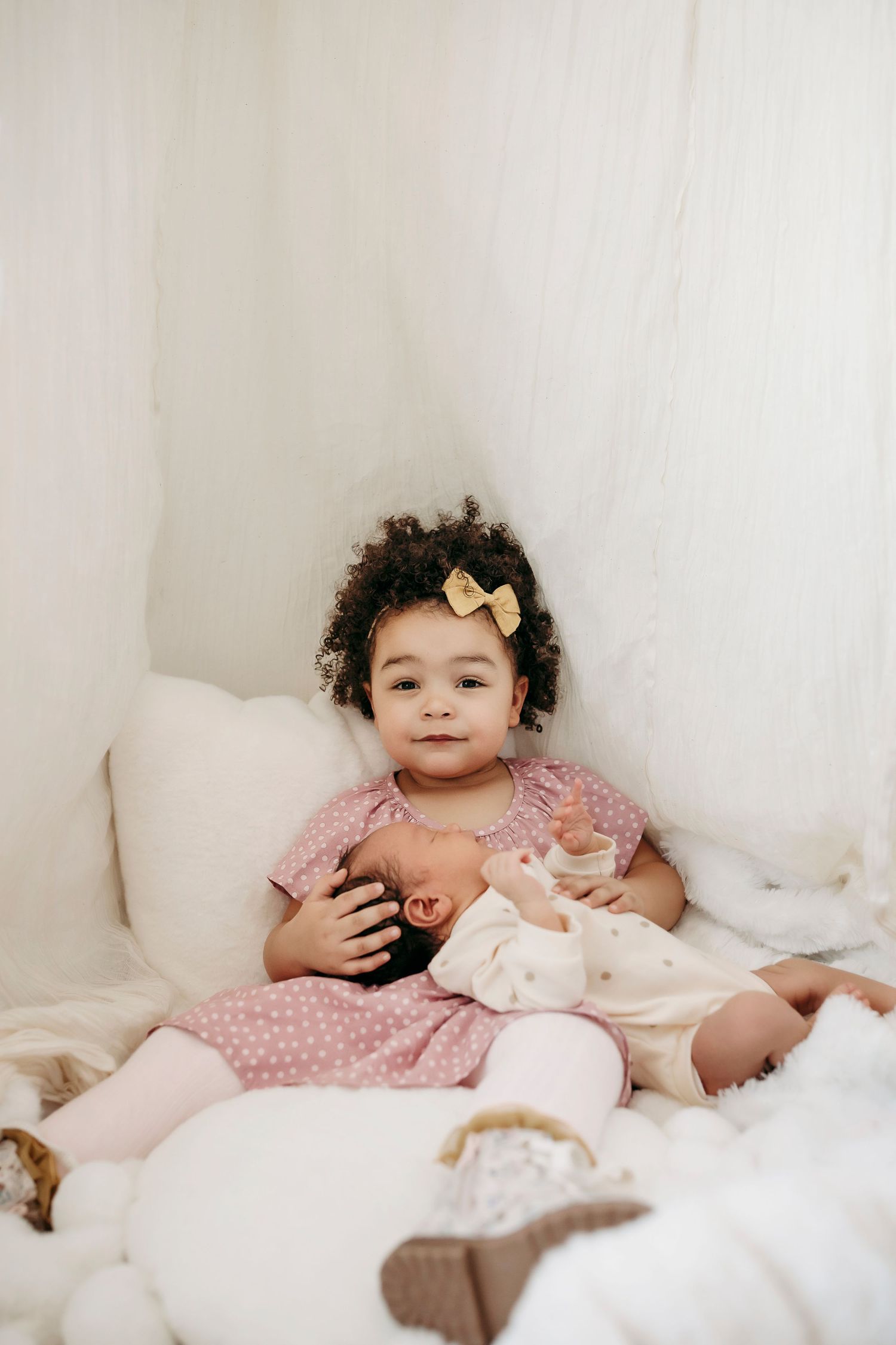 A toddler in a pink polka dot dress sits on white bedding with a yellow bow hair accessory.