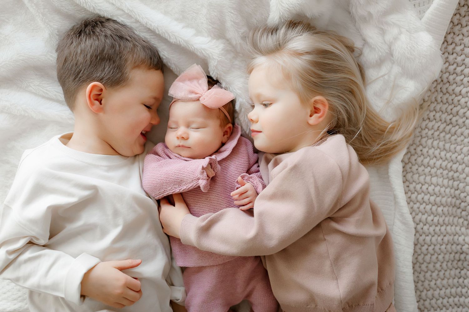 Three young children cuddling together on white bedding.