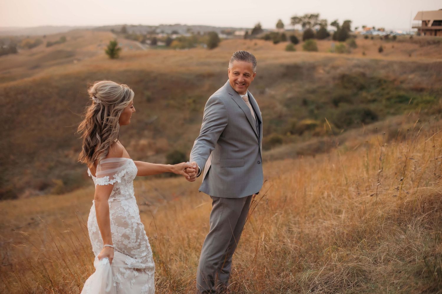 A wedding couple walks hand-in-hand through golden grass on a hillside at sunset.
