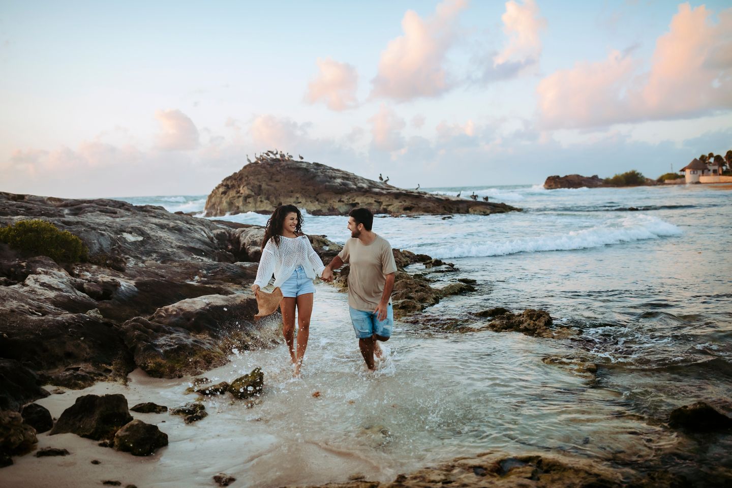 Two people running through shallow water along a rocky beach during sunset.