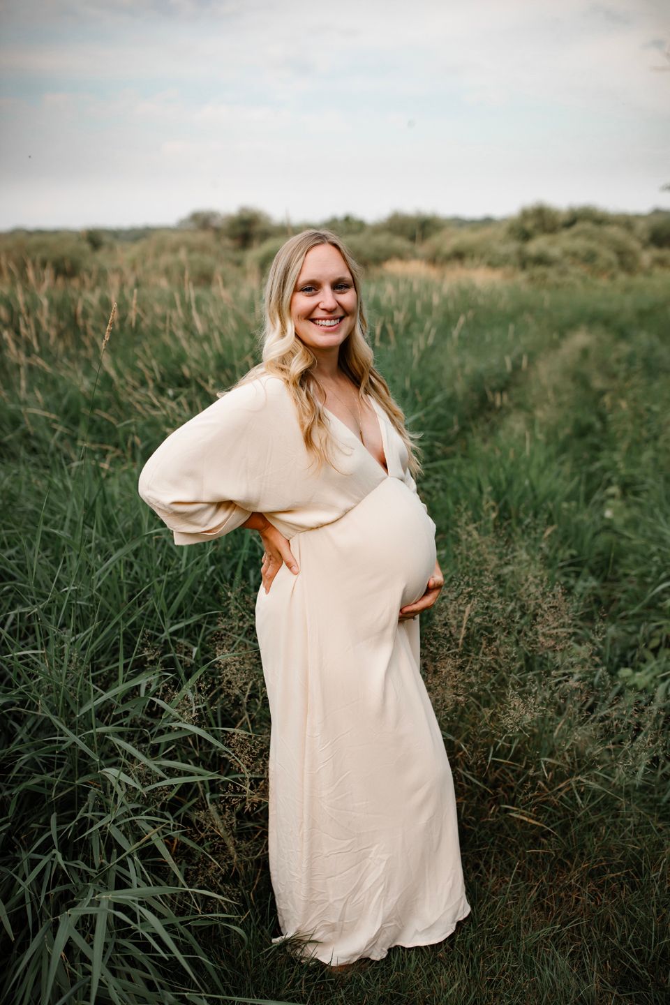 Expectant mother in flowing cream dress poses in tall grass field during sunset maternity photoshoot.