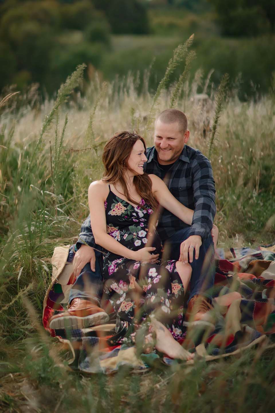 A couple sits together on a blanket in tall grass during a romantic outdoor photoshoot at sunset.