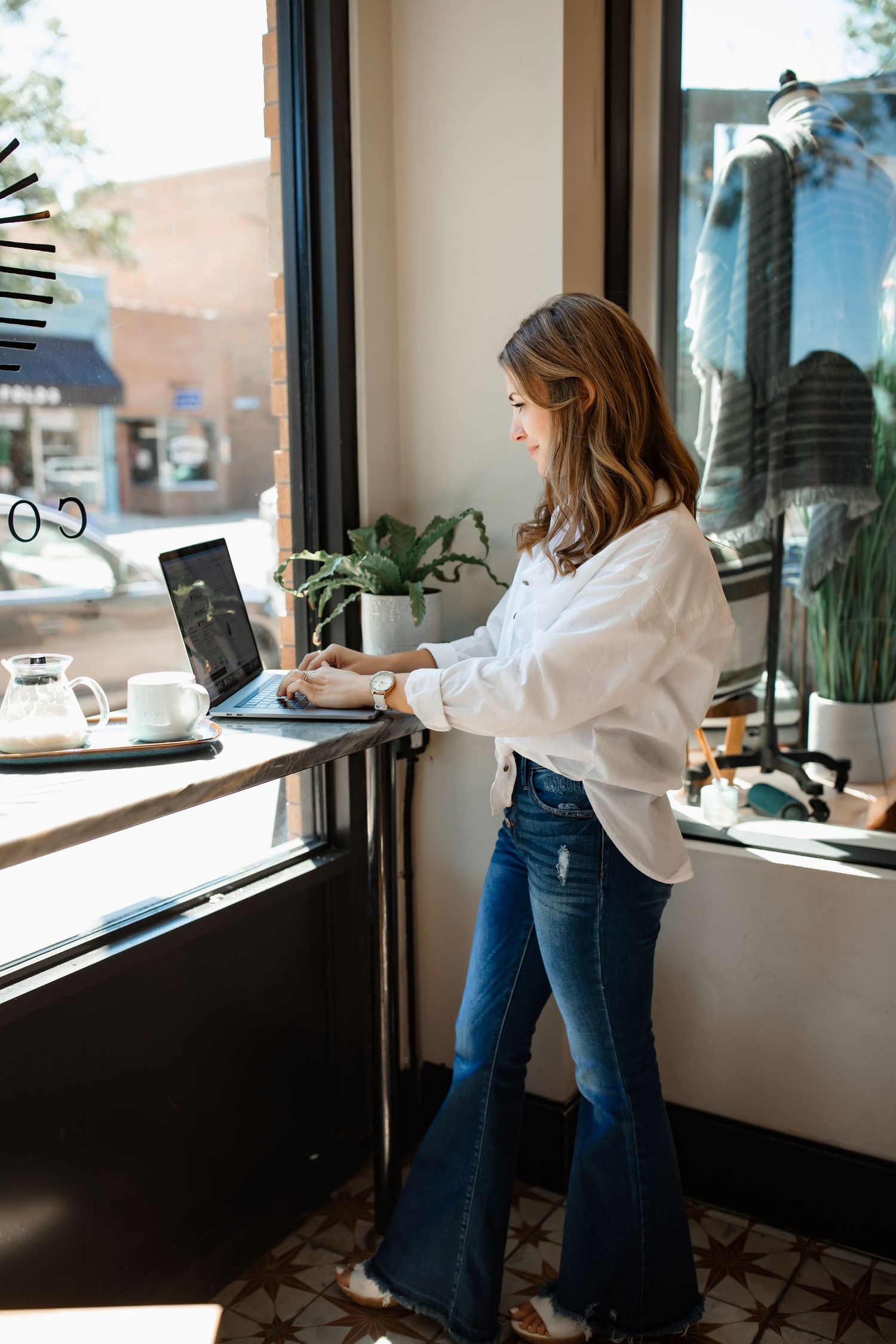 Woman in white shirt and flared jeans working on laptop at counter by window in stylish cafe setting.