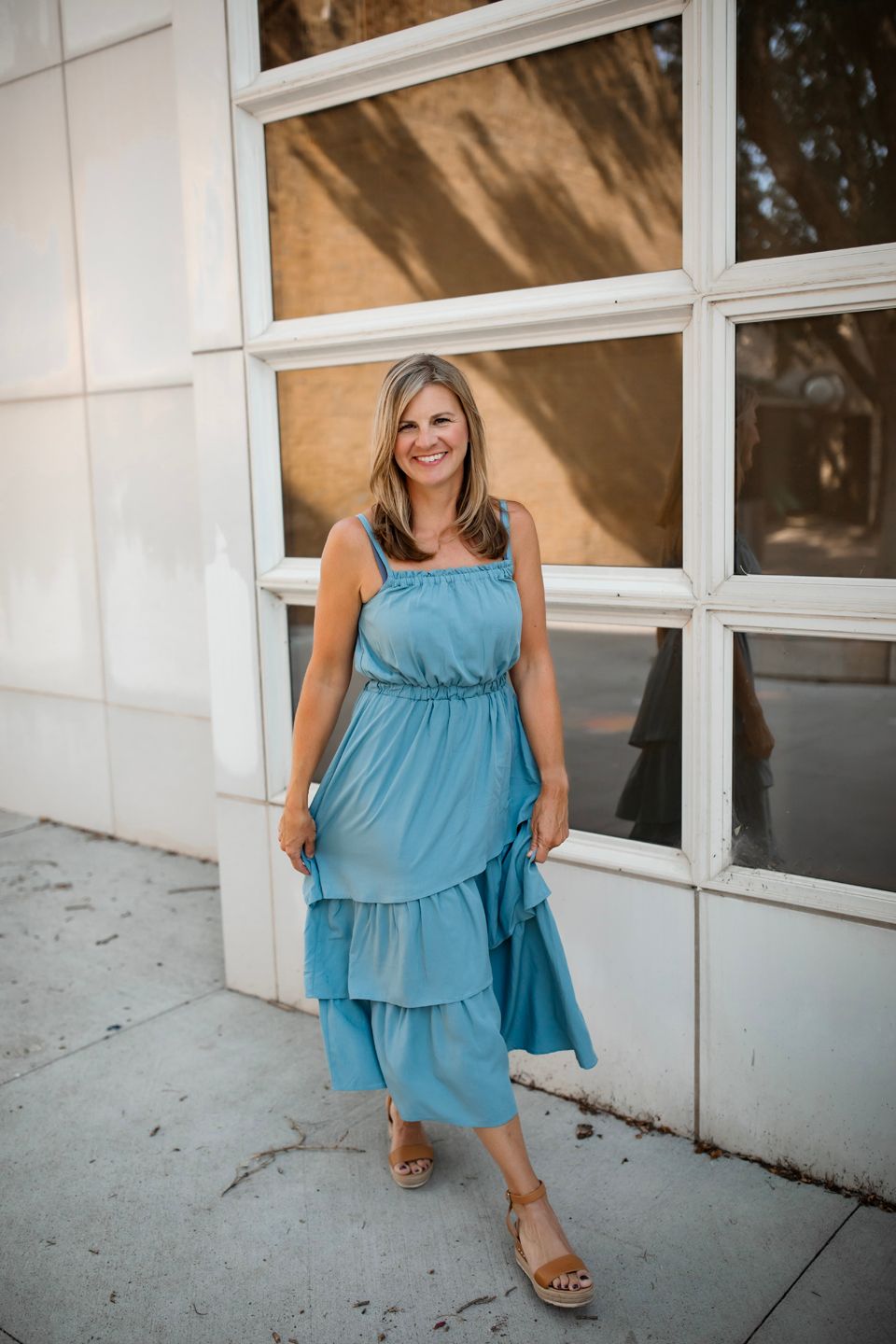 Full length outdoor portrait in a tiered blue midi dress and sandals beside a modern building.