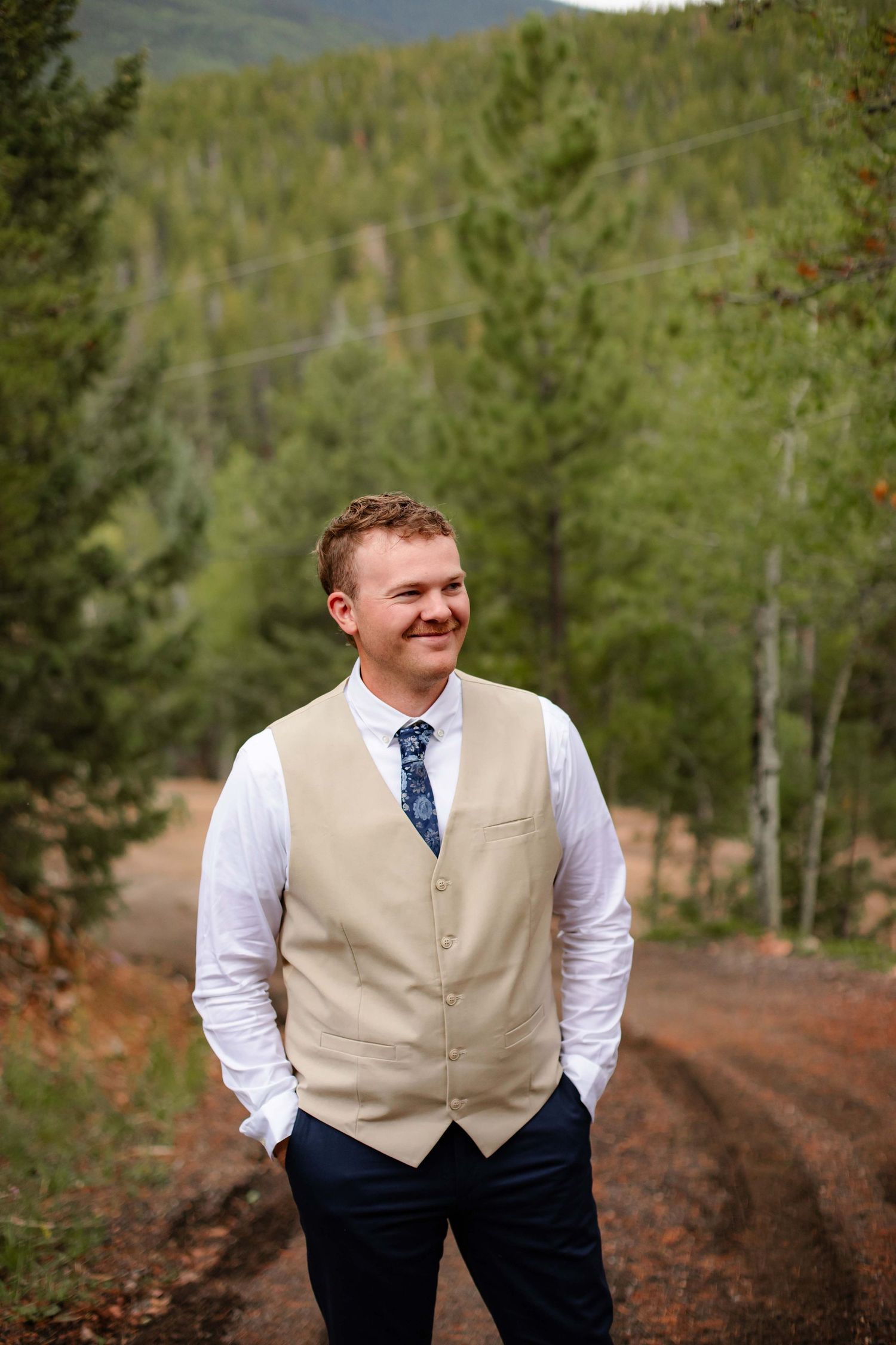 A person in a tan vest and blue tie stands outdoors against a forested mountain backdrop.