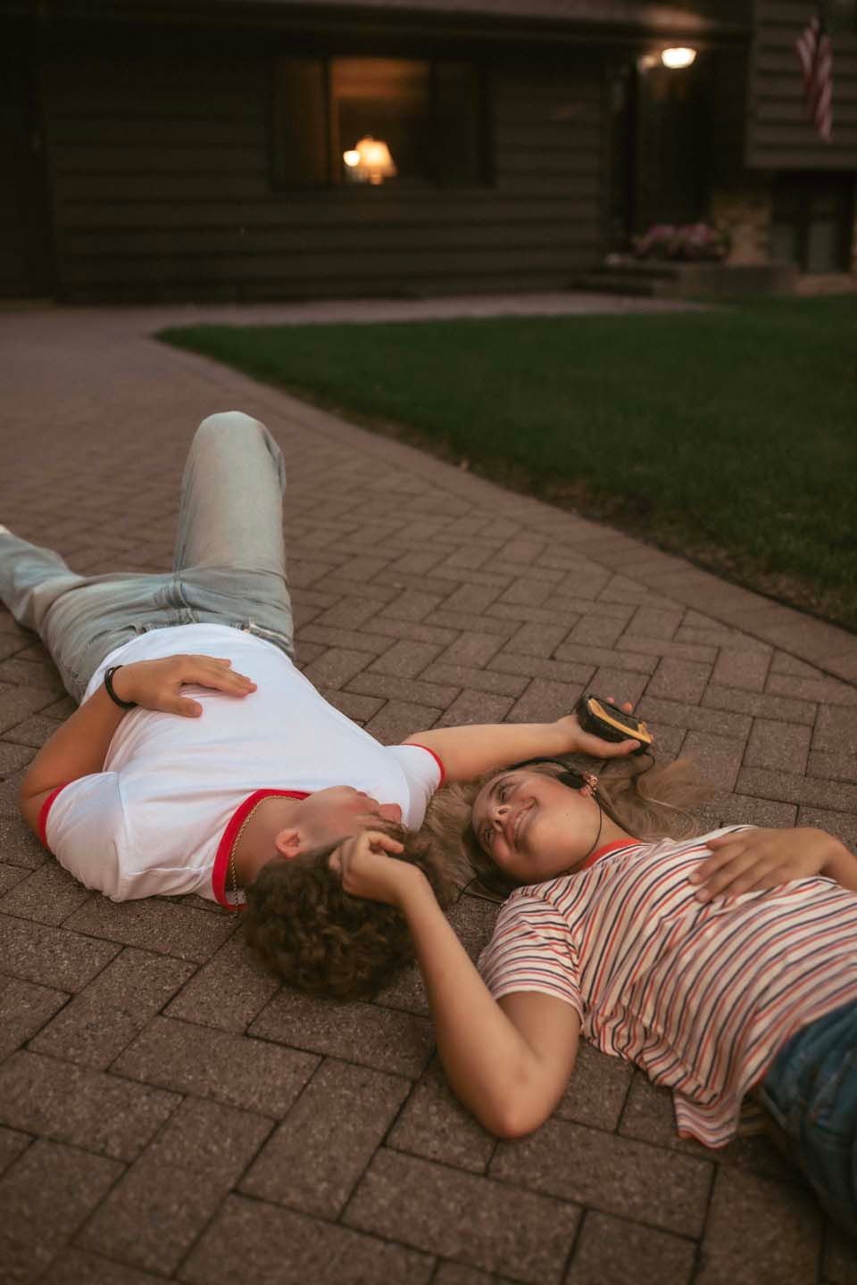 Young couple lying on a brick pathway at dusk, laughing and holding a phone outside a house.