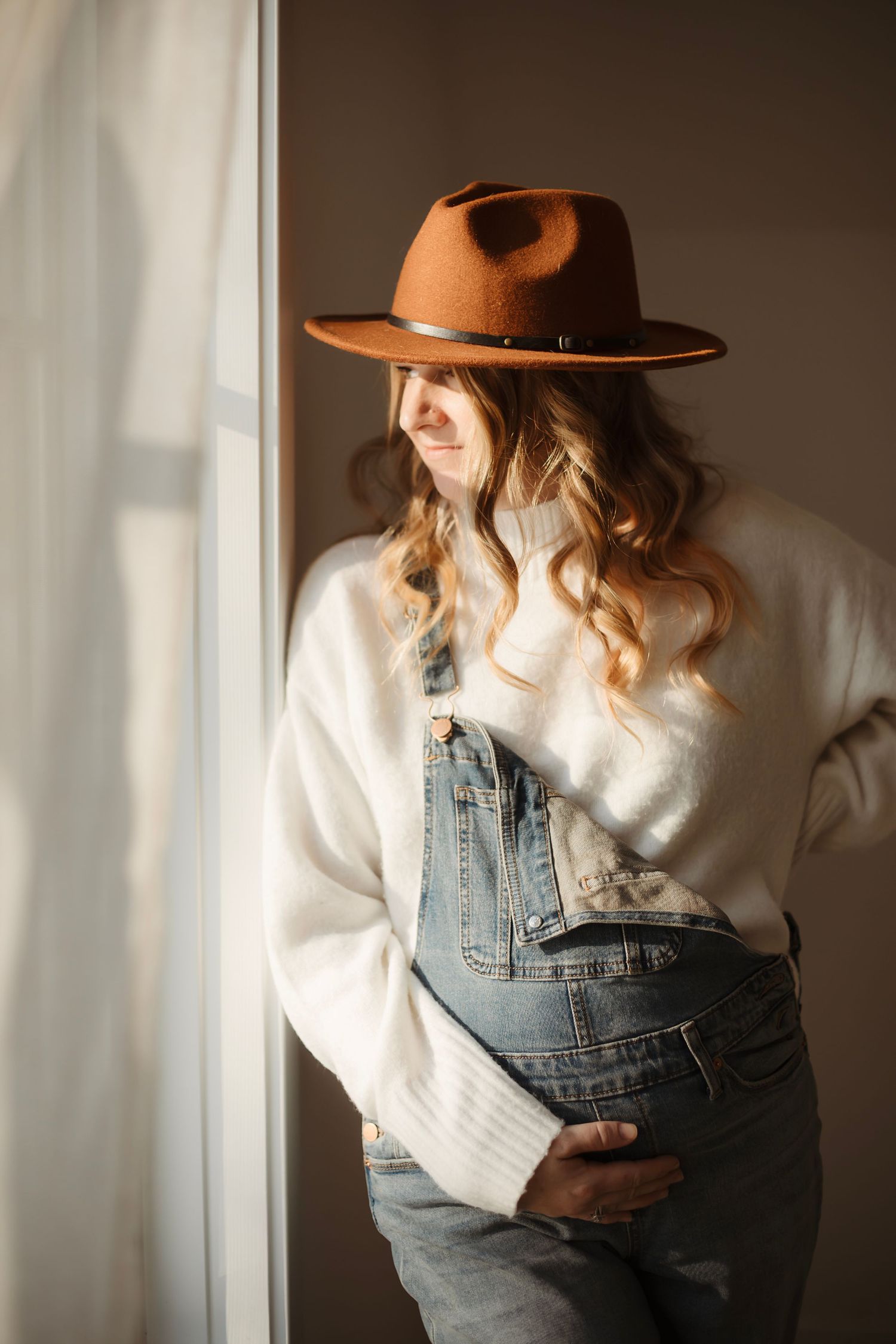 Pregnant woman in white sweater, denim overalls, and brown felt hat standing by a sunlit window. Willow & Wren Photography maternity photo s