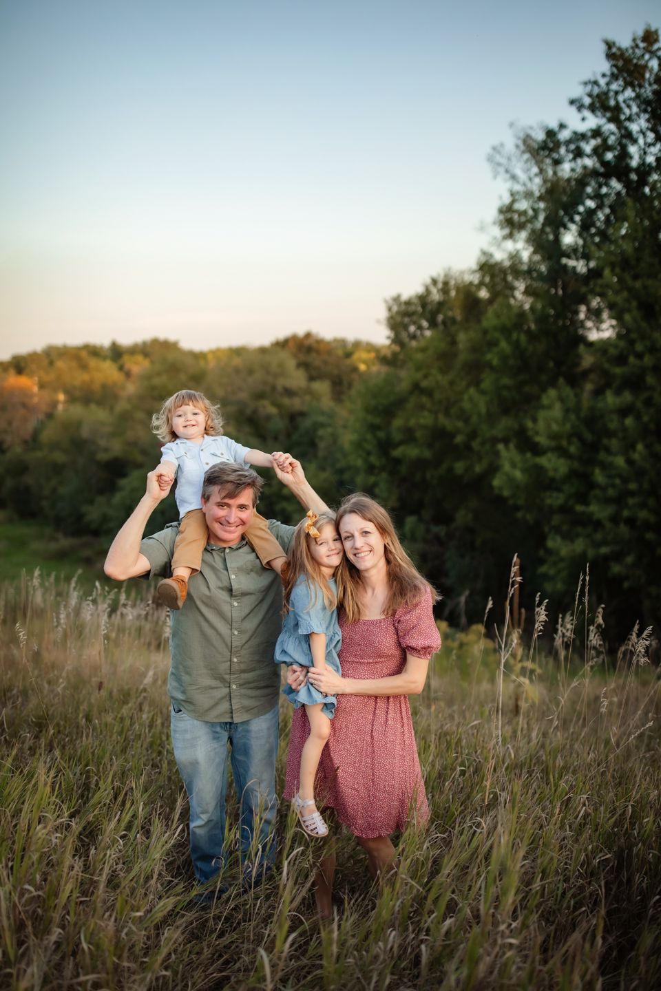 Family poses together in tall grass meadow at sunset with warm golden lighting and natural outdoors backdrop.