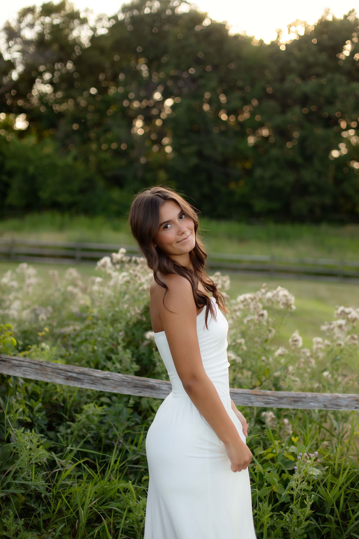 Young woman in a white strapless dress standing in a sunlit field with wildflowers and a wooden fence.