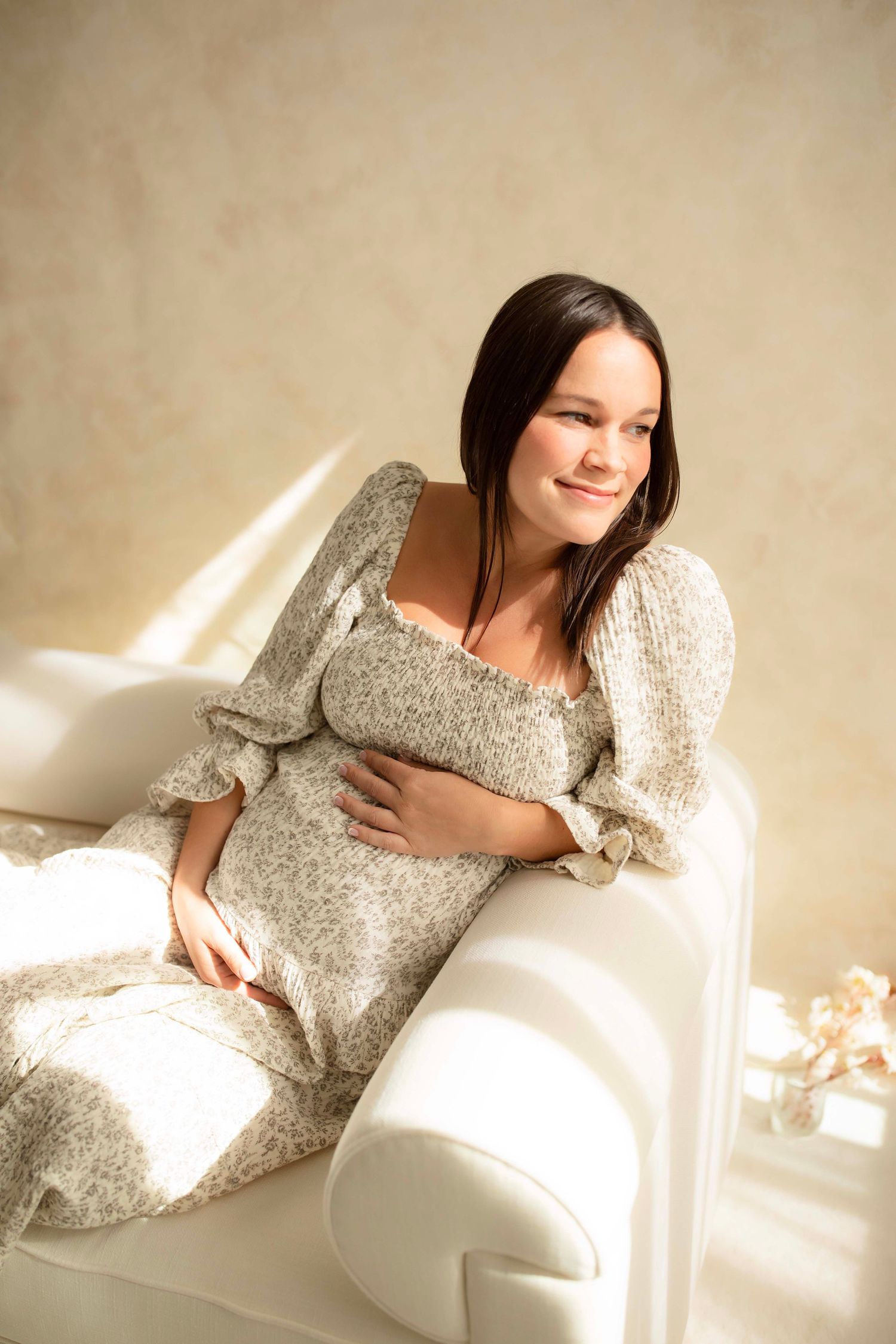 Pregnant woman in floral dress sitting on white sofa, gently cradling her baby bump.
