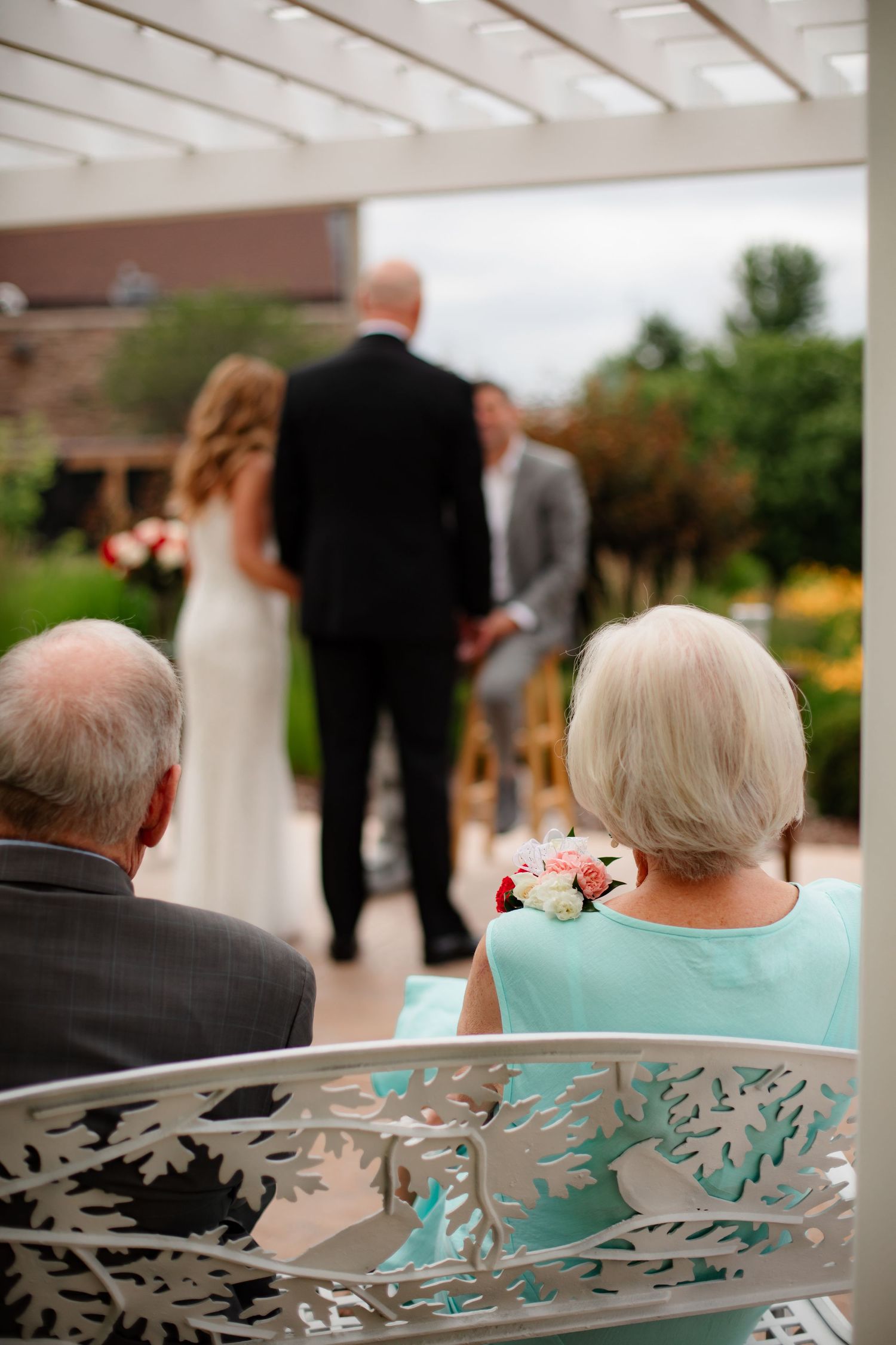 Wedding guests seated in ornate chairs with decorative metalwork backs at an outdoor ceremony venue.