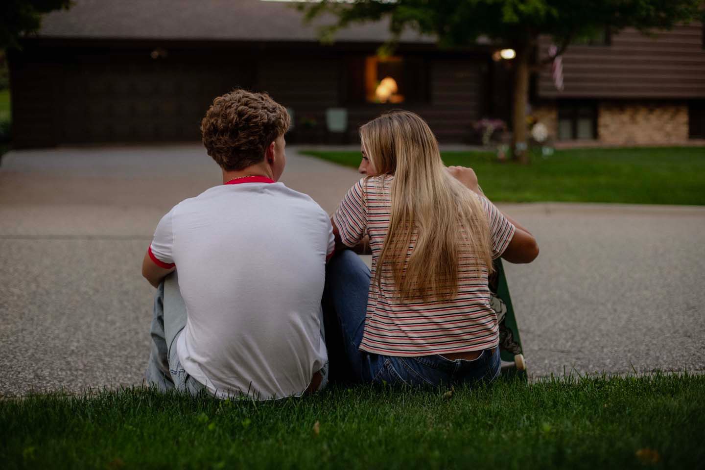 A young couple sits together on green grass, viewed from behind, near a building at dusk.