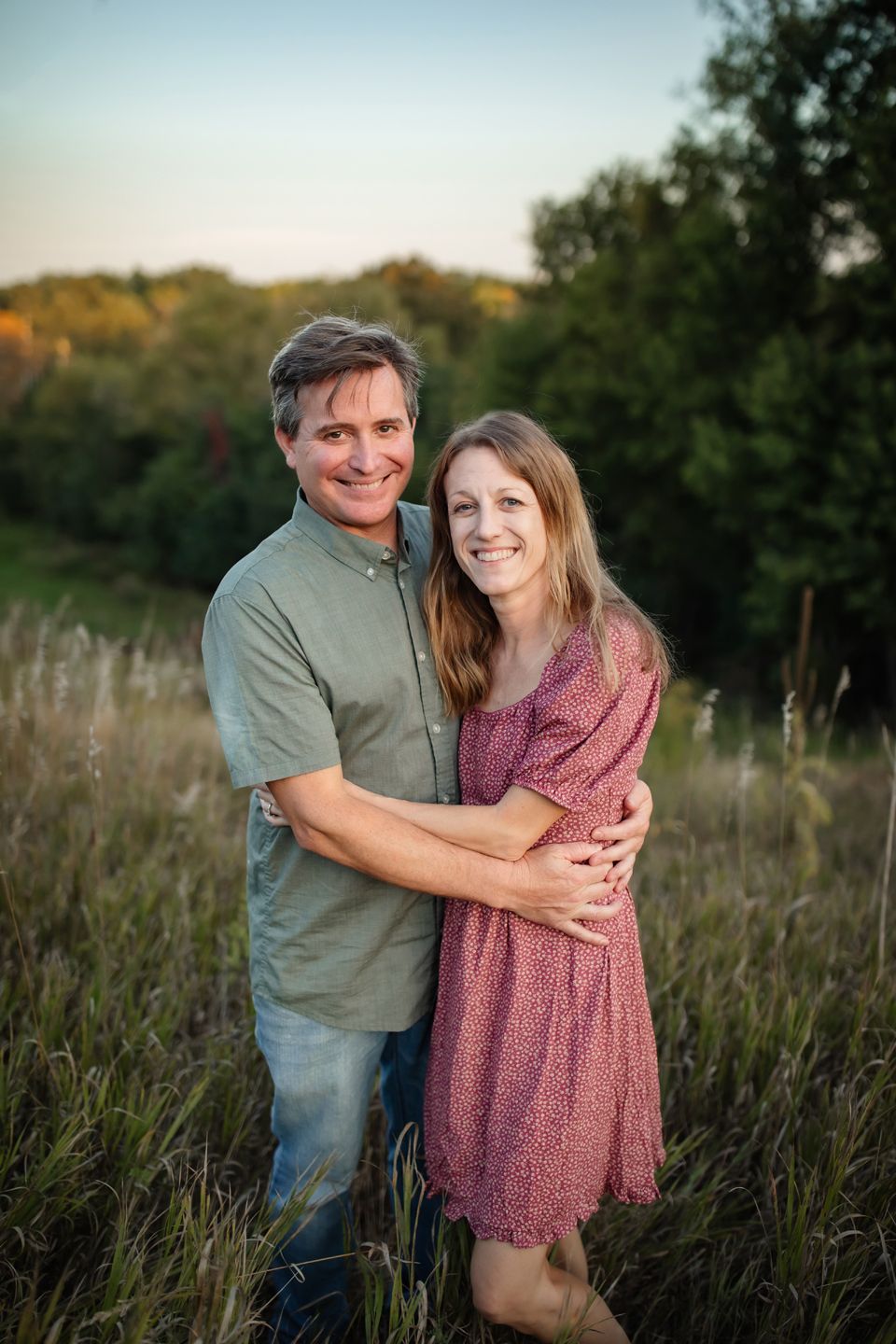 A couple embraces in a sunlit meadow during golden hour with tall grass and trees in the background.