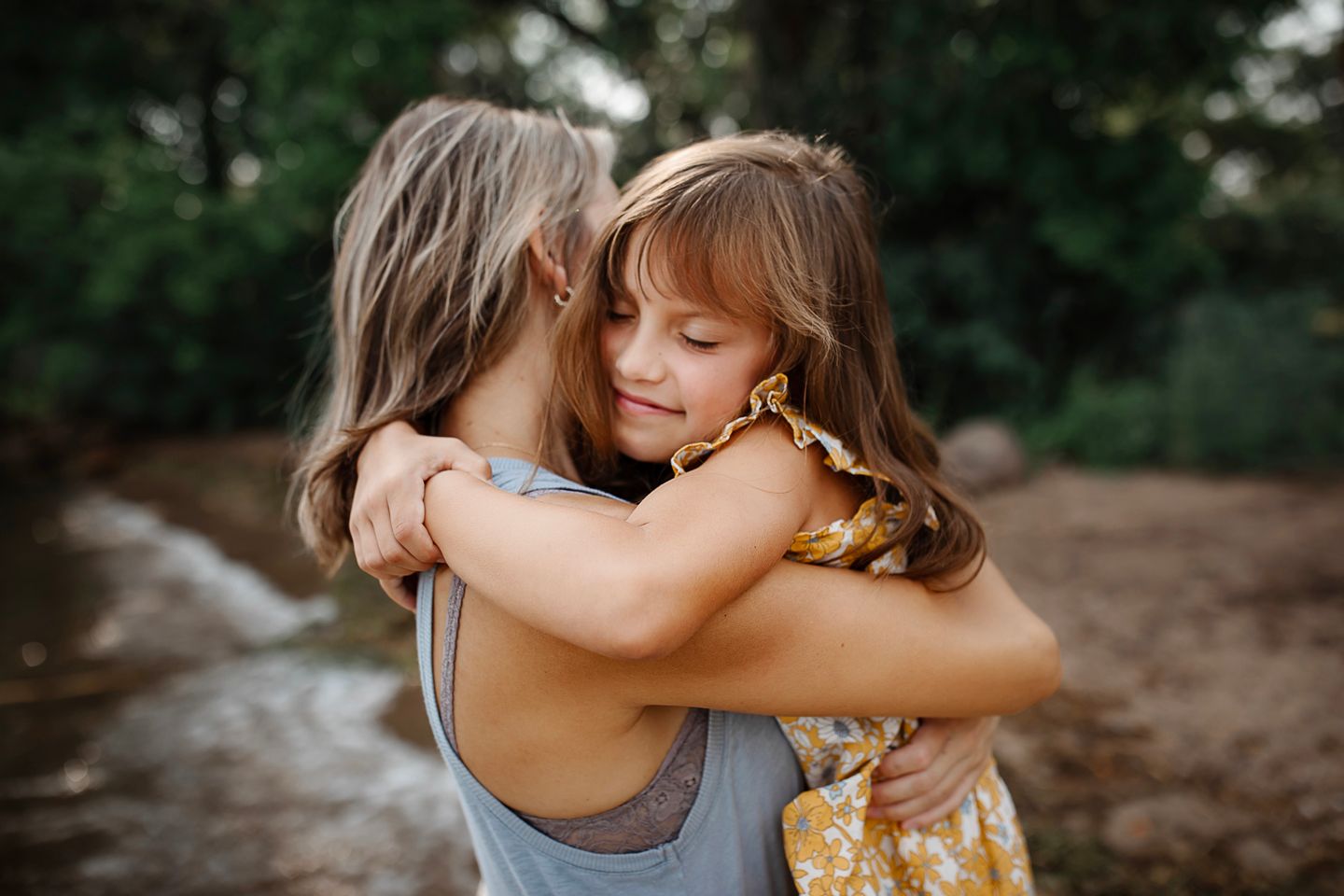 Two figures embrace in a tender hug outdoors during golden hour with a natural backdrop of trees and earth.
