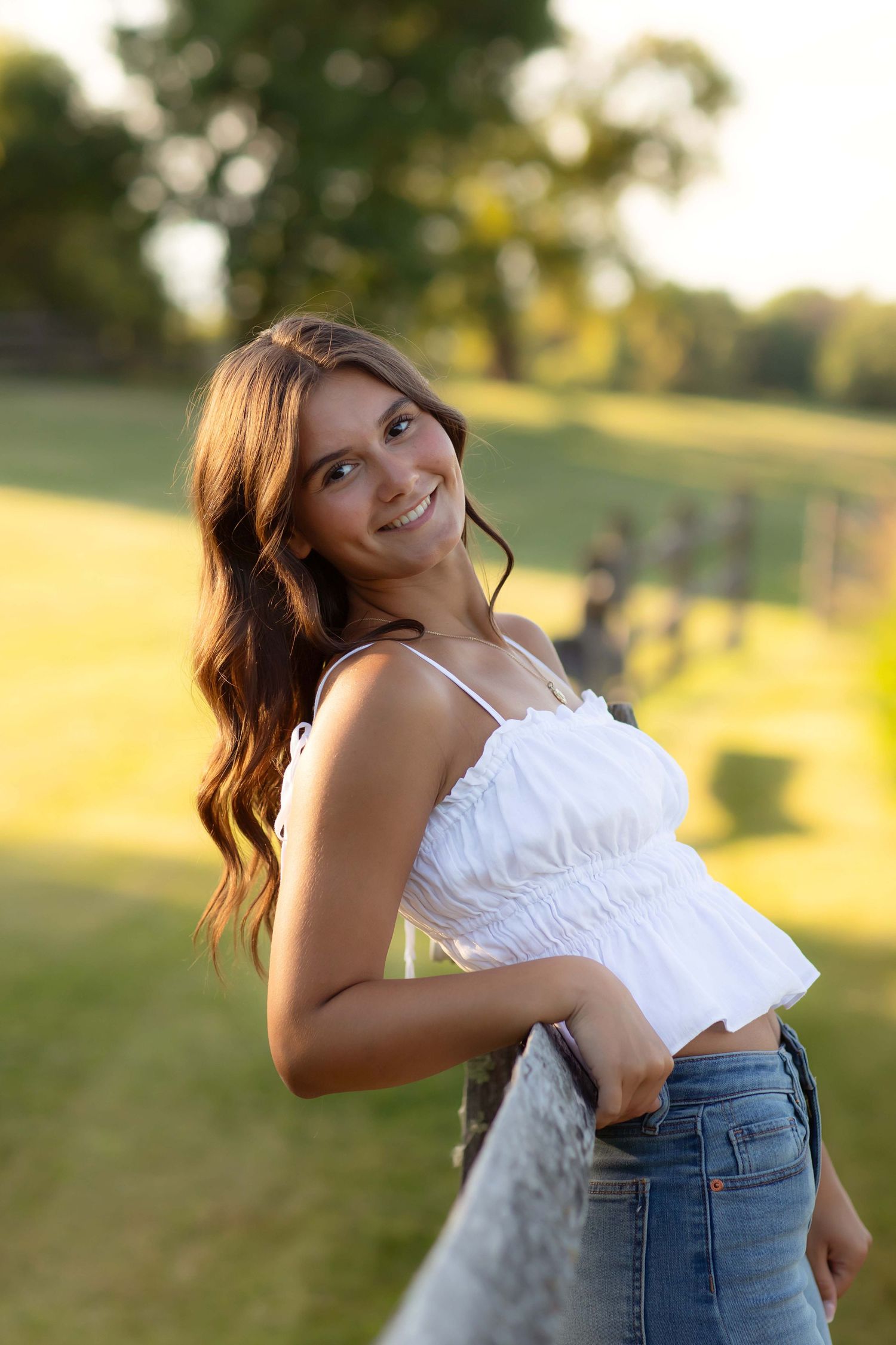 Smiling young woman in white crop top and jeans posing outdoors in a sunny green park setting.