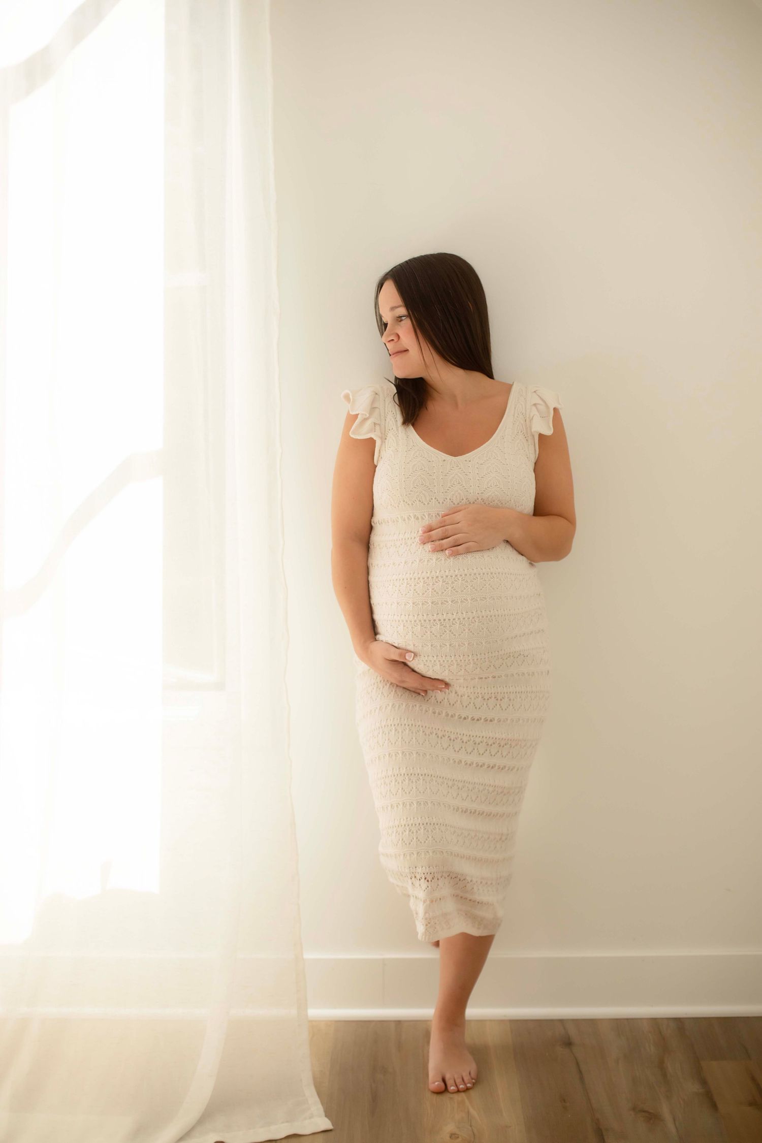 Pregnant woman in white lace dress cradling baby bump, leaning against wall near sunlit window.
