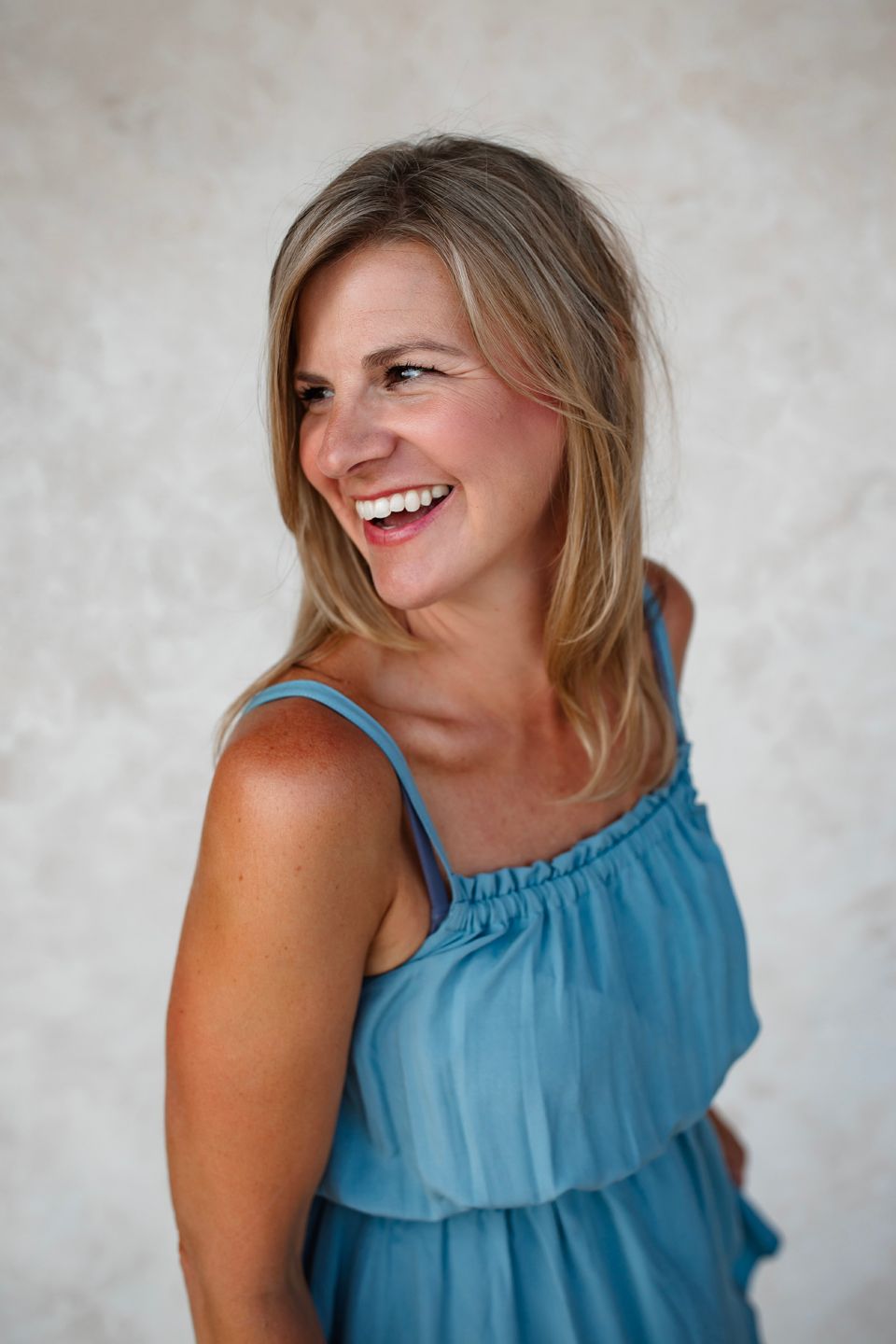 Candid laughing portrait in a flowing blue sleeveless dress against a light wall.