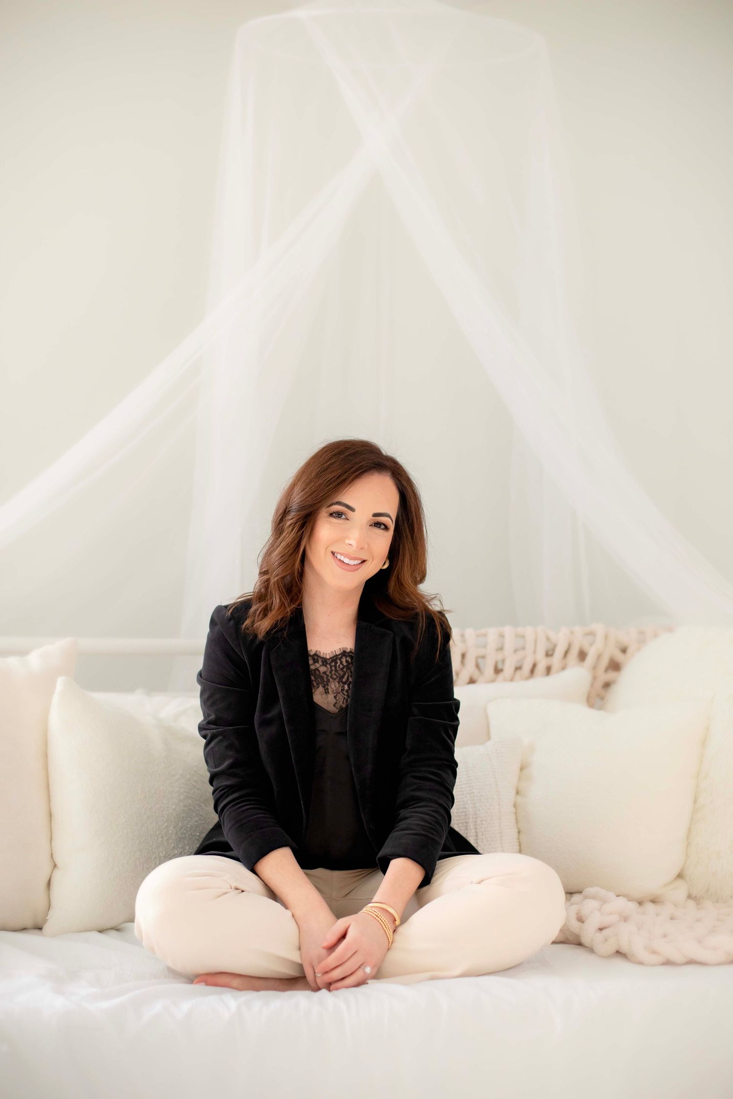 Relaxed indoor portrait in black blazer sitting cross-legged on white couch with draping.
