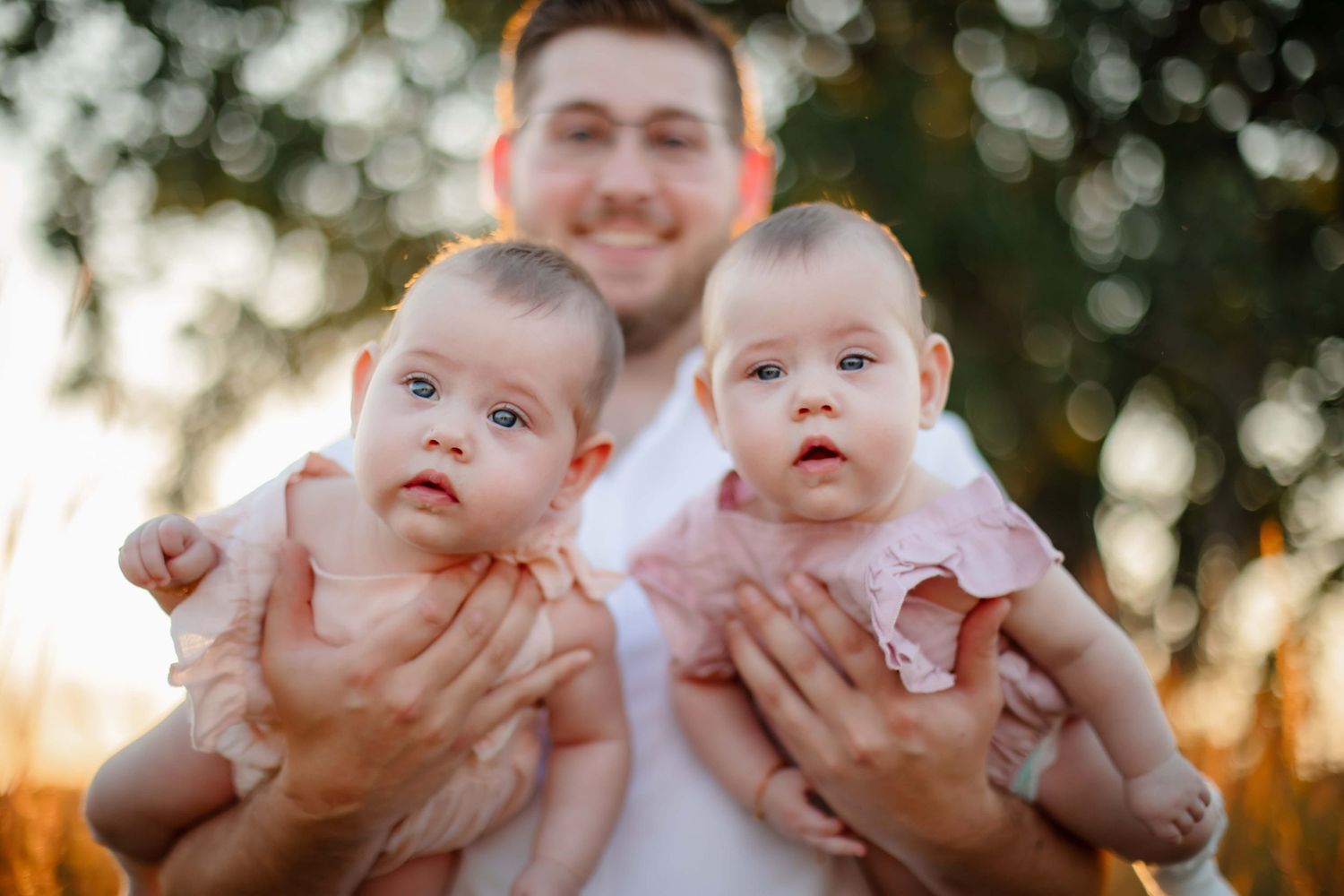 Two small babies being held up against a bokeh background at sunset.