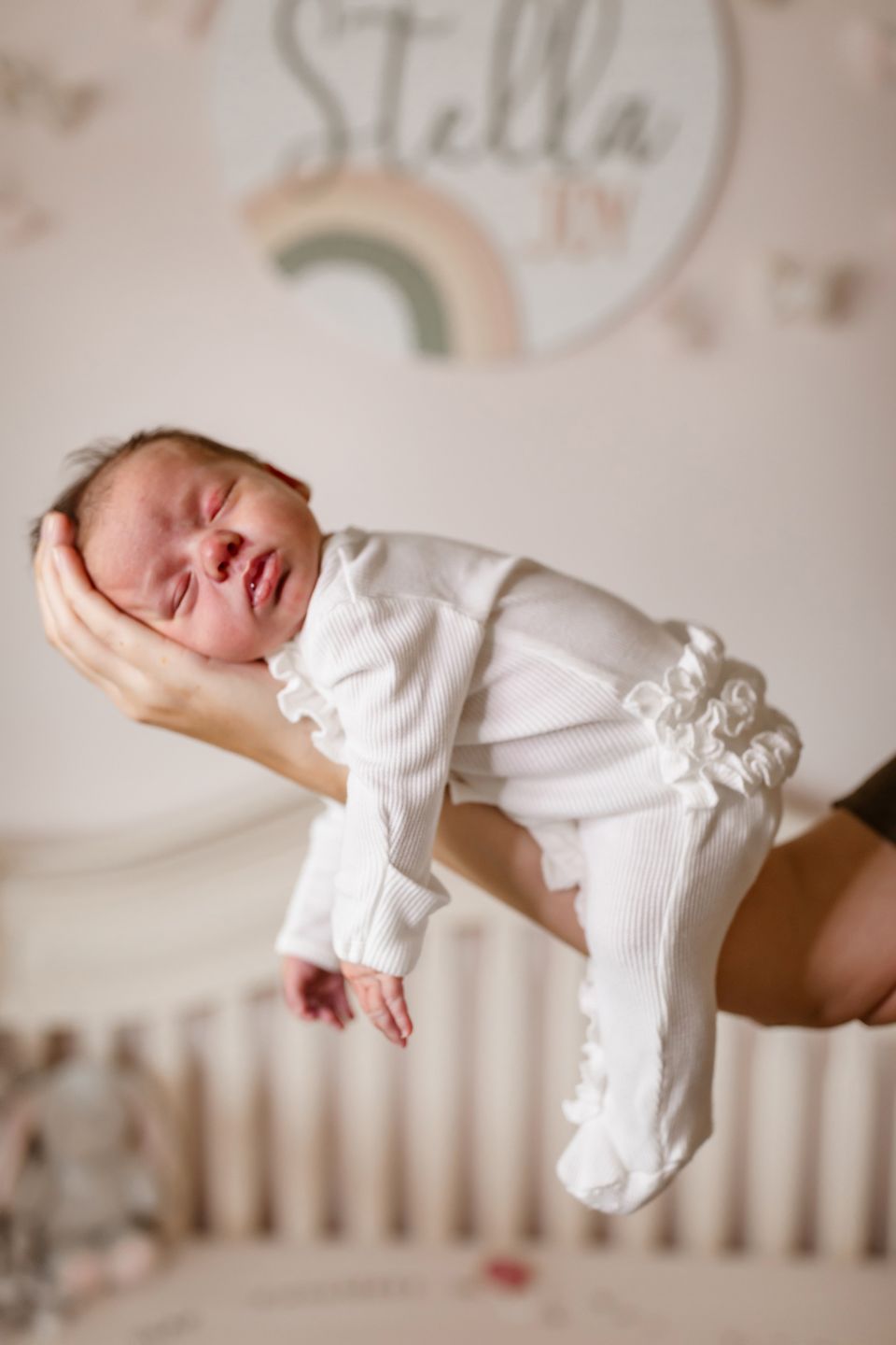 A sleeping newborn in white ruffled sleepwear held gently against a pastel nursery backdrop.