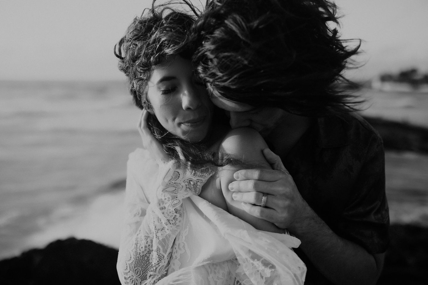 Black and white photo of intimate beachside embrace with wind-swept hair.