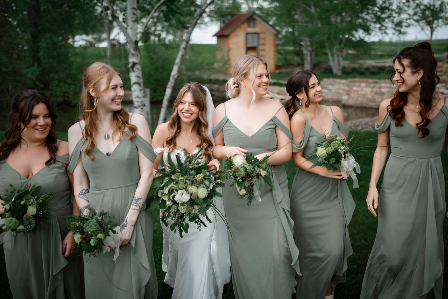 Bridesmaids in sage green dresses share candid moment with bride outdoors.