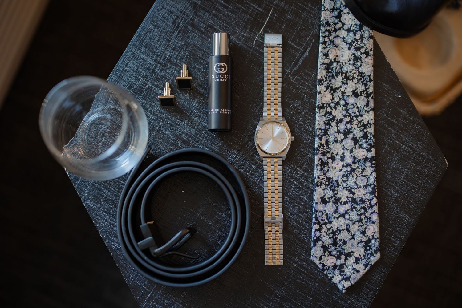 Groom's accessories including watch, belt, tie and cufflinks arranged on dark surface.