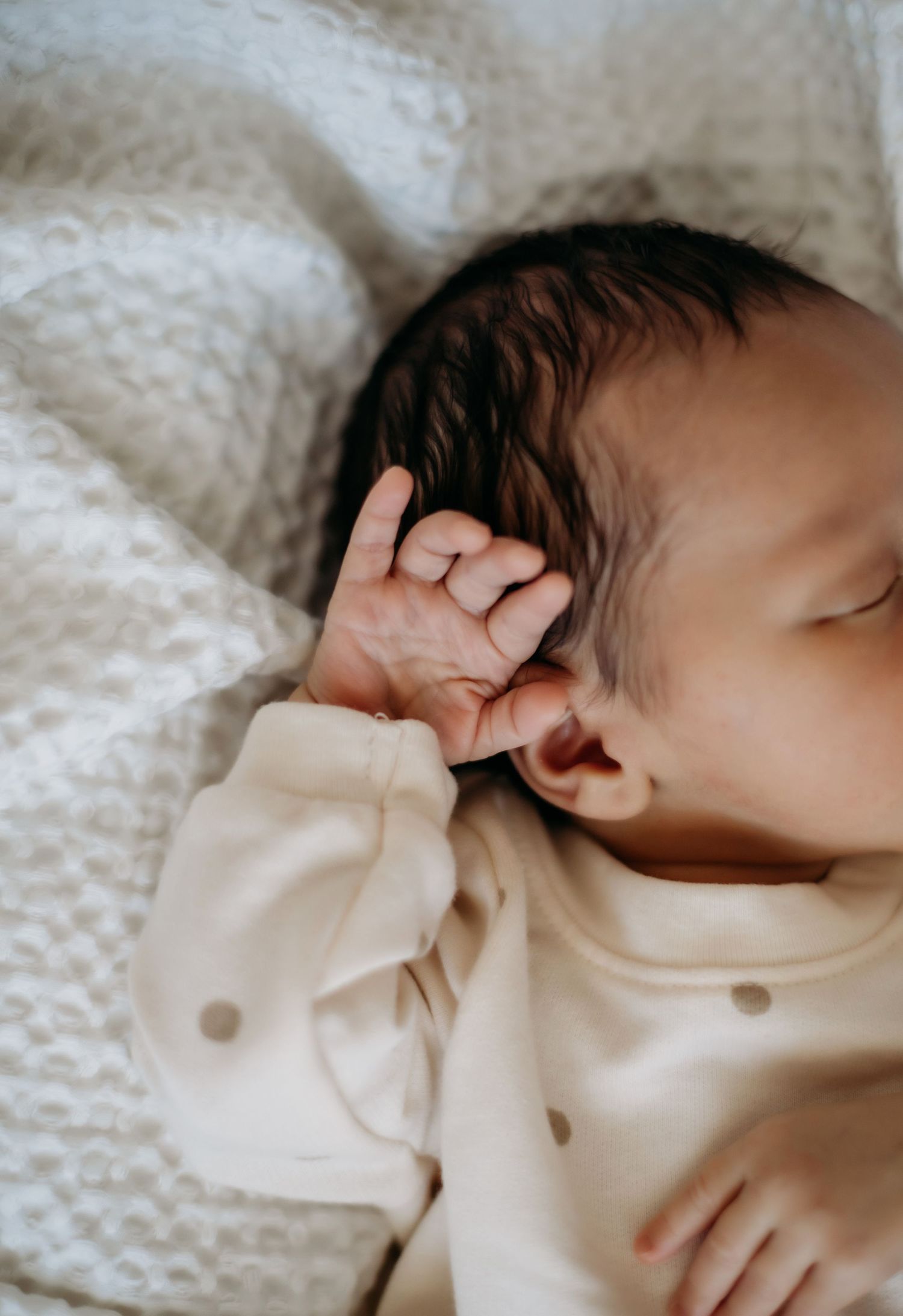 Close-up of sleeping baby in cream polka dot outfit with hand near face on white blanket.
