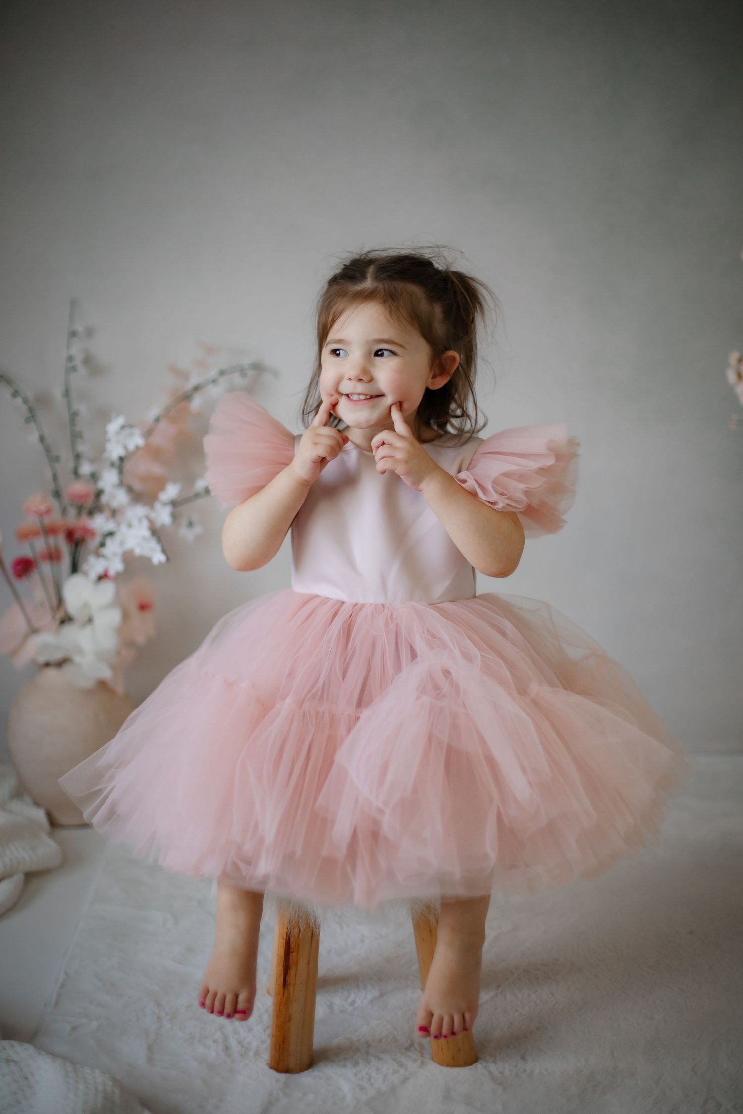Child in pink tulle dress poses playfully on wooden stool with floral backdrop.