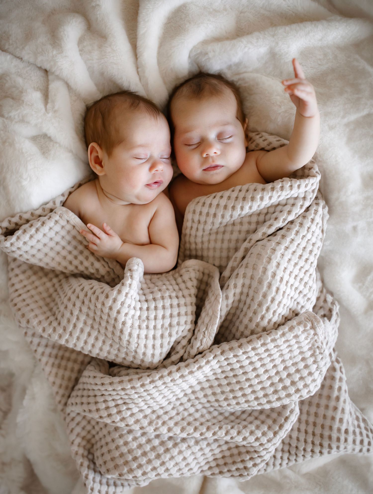 Two sleeping infants cuddled together under textured waffle blanket on white bedding.