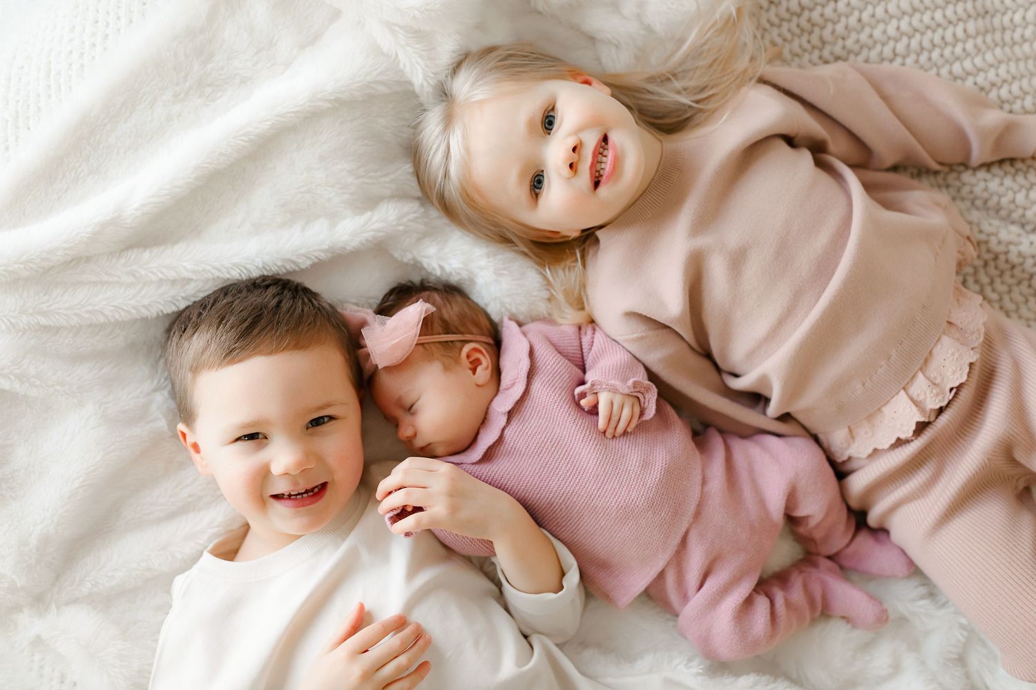 Three siblings lying together on white bedding with joyful expressions.