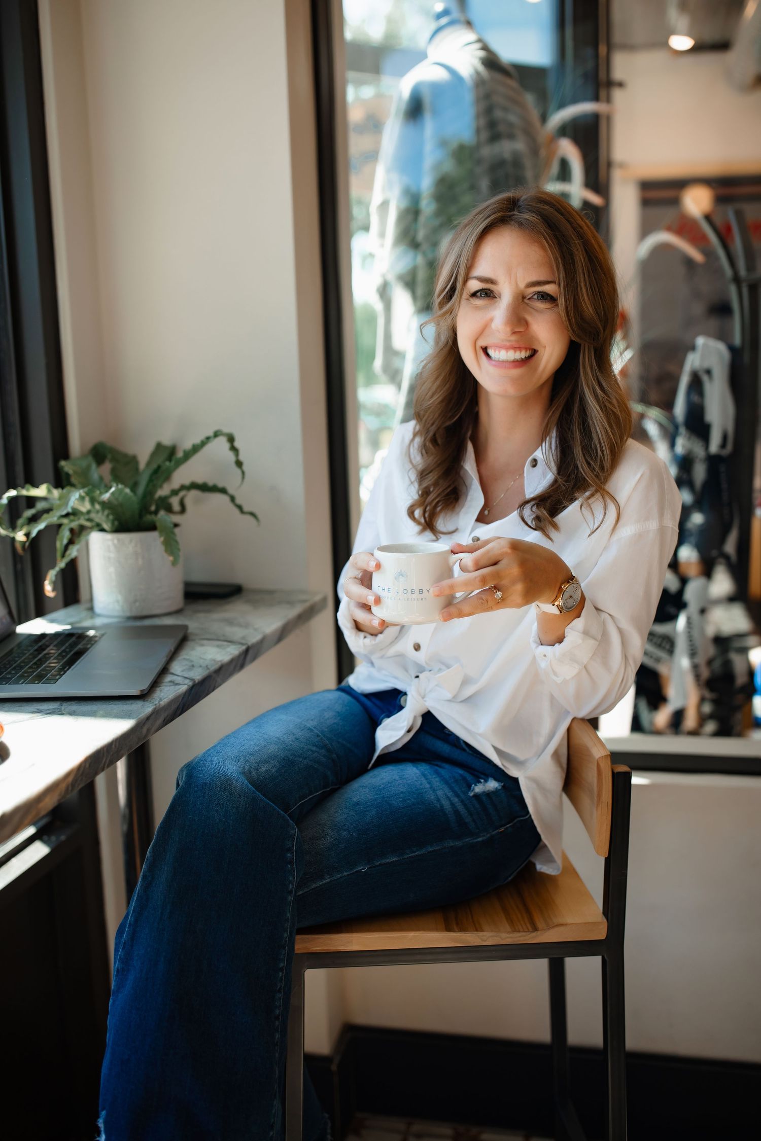Casual coffee shop portrait of someone in white shirt holding ceramic mug by window.