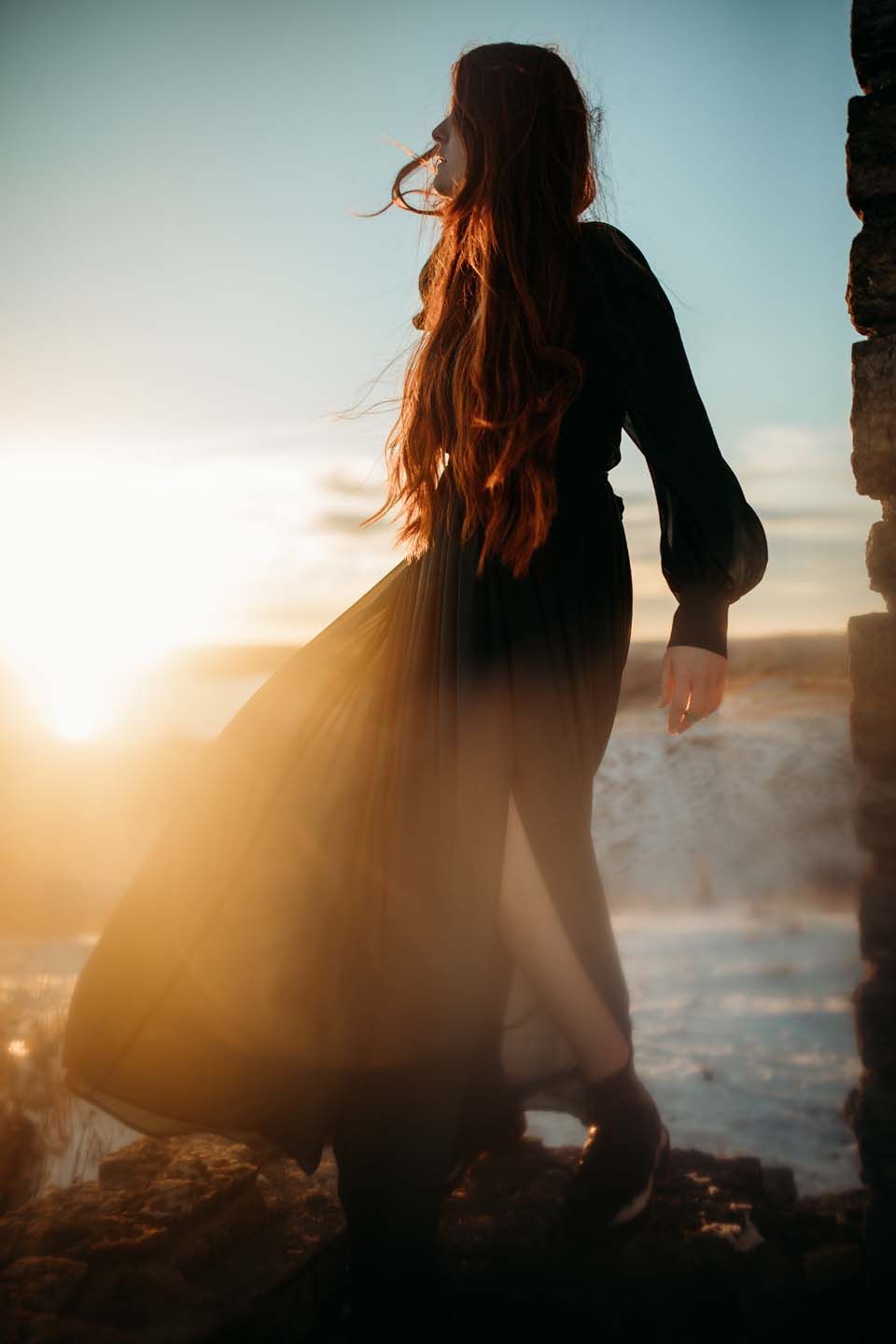 Silhouette of person with flowing dress and long hair against seaside sunset.