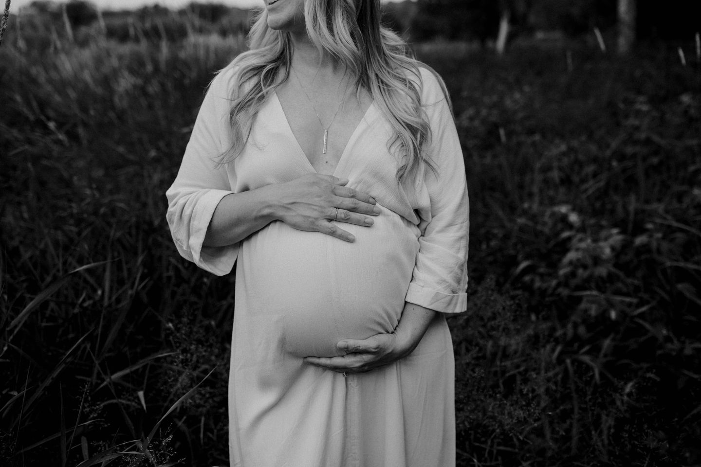 A pregnant figure in a white dress holds their belly while standing in tall grass, captured in black and white.