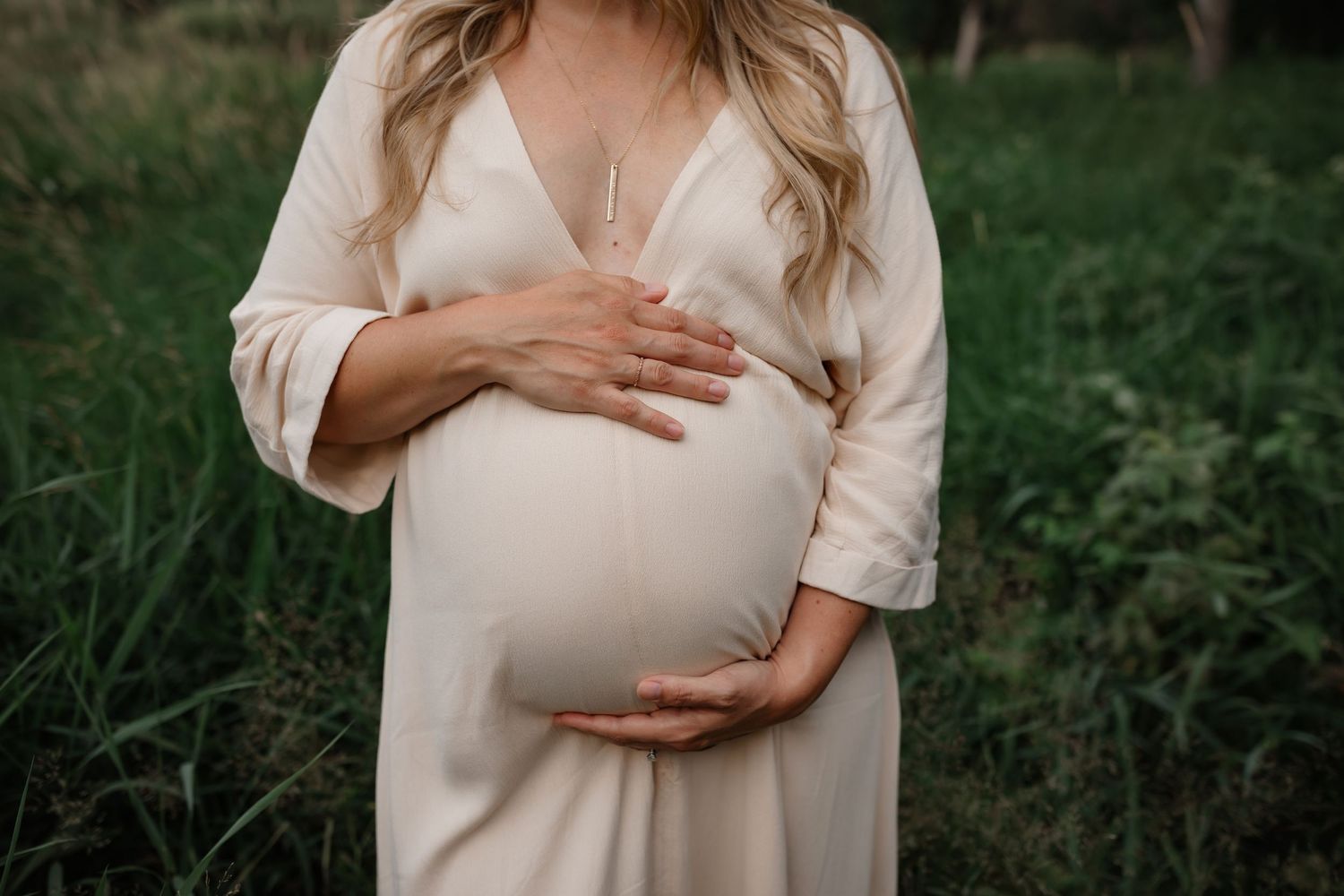 Expecting mother in cream dress holds her baby bump while standing in grassy outdoor setting in natural light.