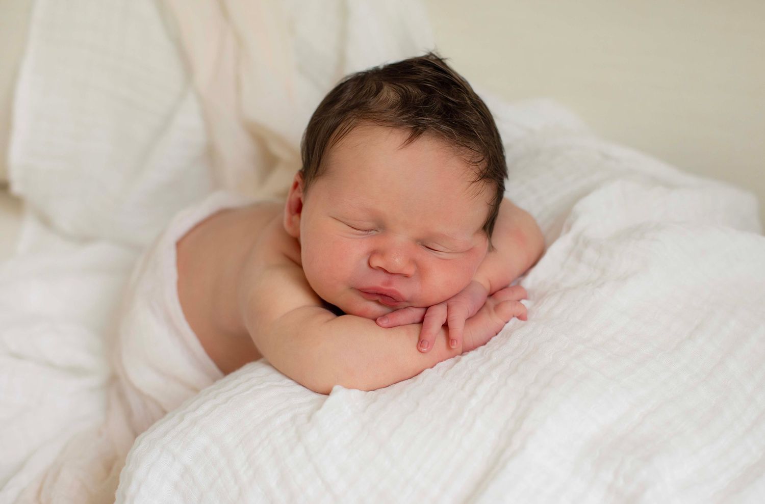 Peaceful newborn baby with dark hair sleeping on a soft white blanket, resting chin on tiny hands.