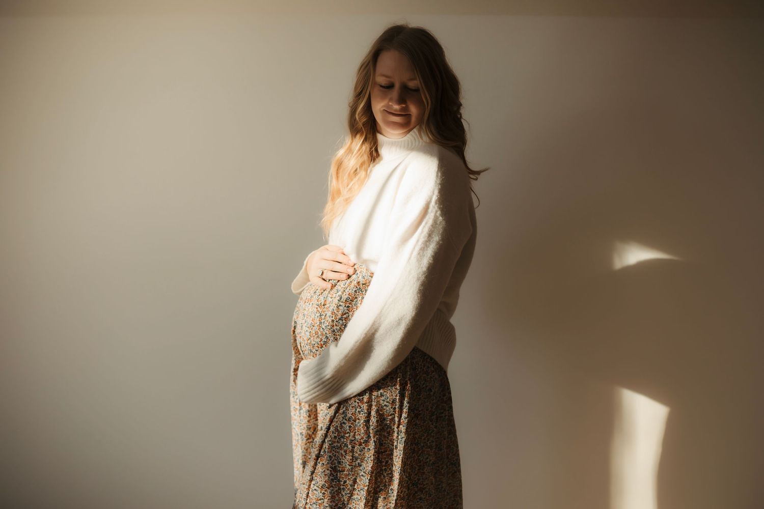 Pregnant woman in white sweater and floral skirt cradling baby bump in soft natural light. Willow & Wren Photography maternity photo session