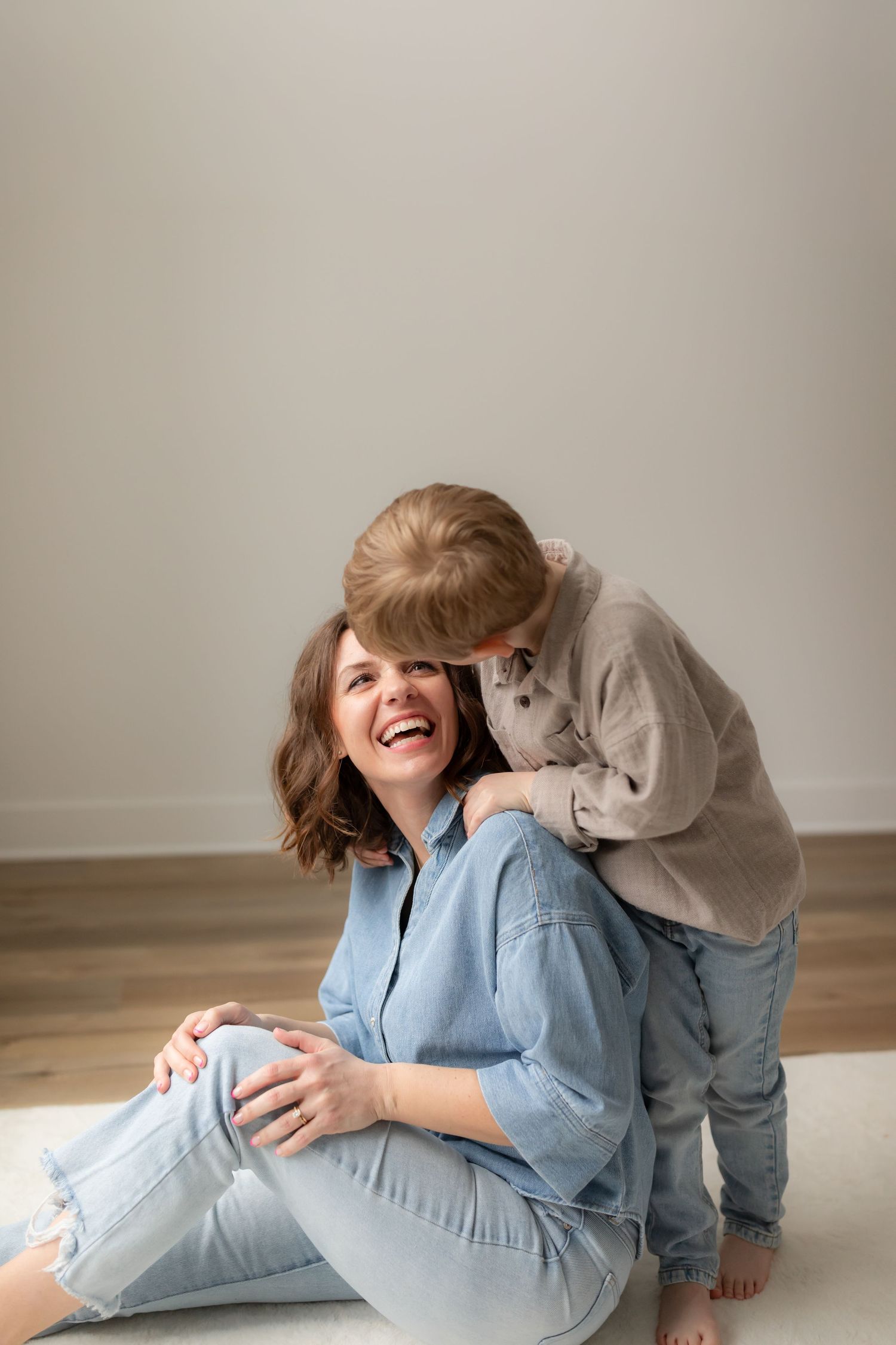 Parent and child play and laugh together on wooden floor during casual indoor photoshoot.