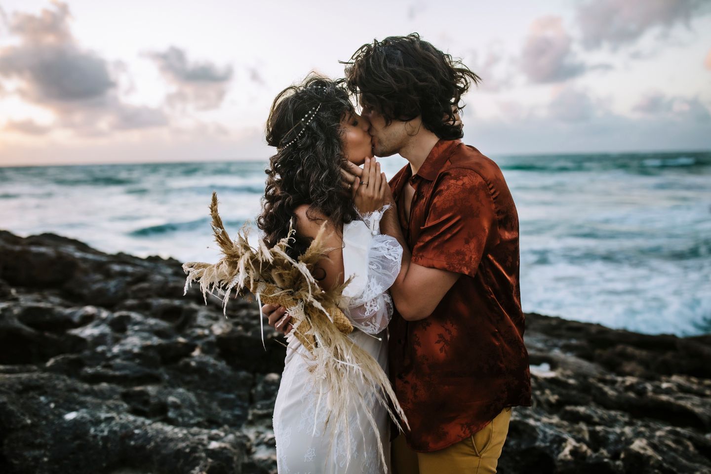 A couple shares an intimate moment with pampas grass bouquet against dramatic ocean backdrop.