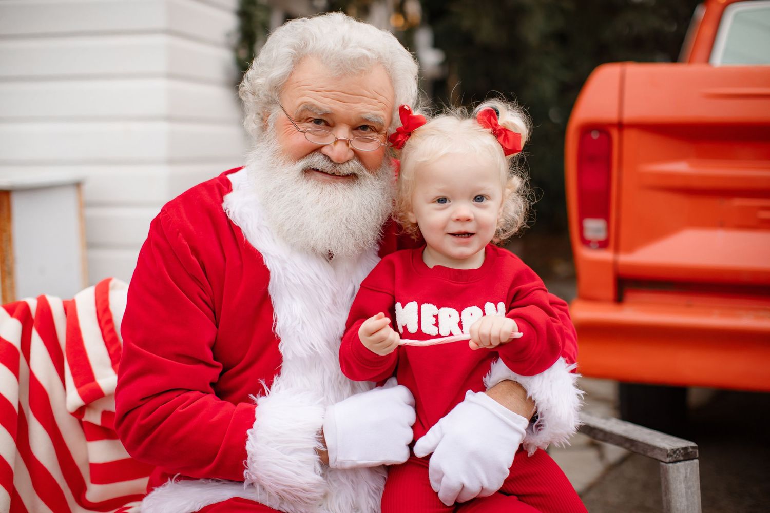 Santa Claus sitting with small child in matching red outfits near vintage red truck.