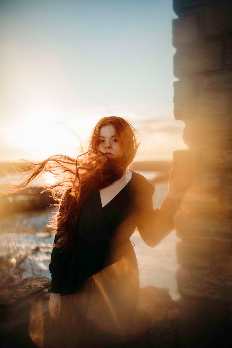 A dramatic sunset silhouette against stone ruins with flowing fabric and hair in the wind.