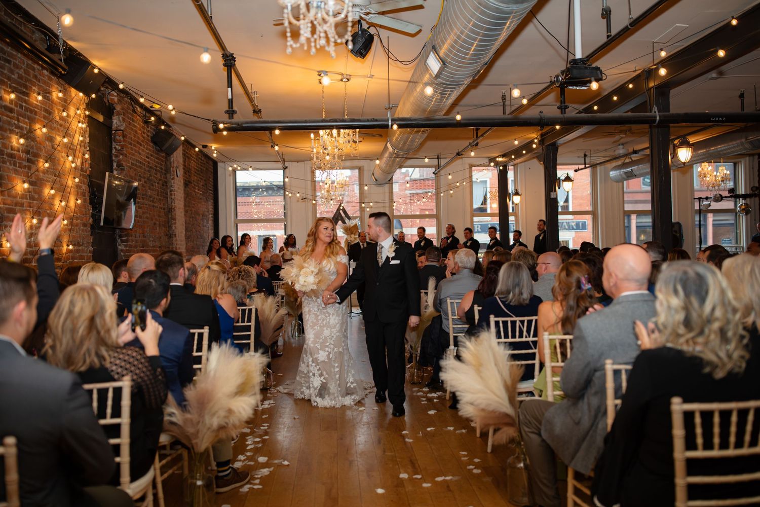 Indoor wedding ceremony in an industrial-style venue with exposed brick walls and twinkling lights.