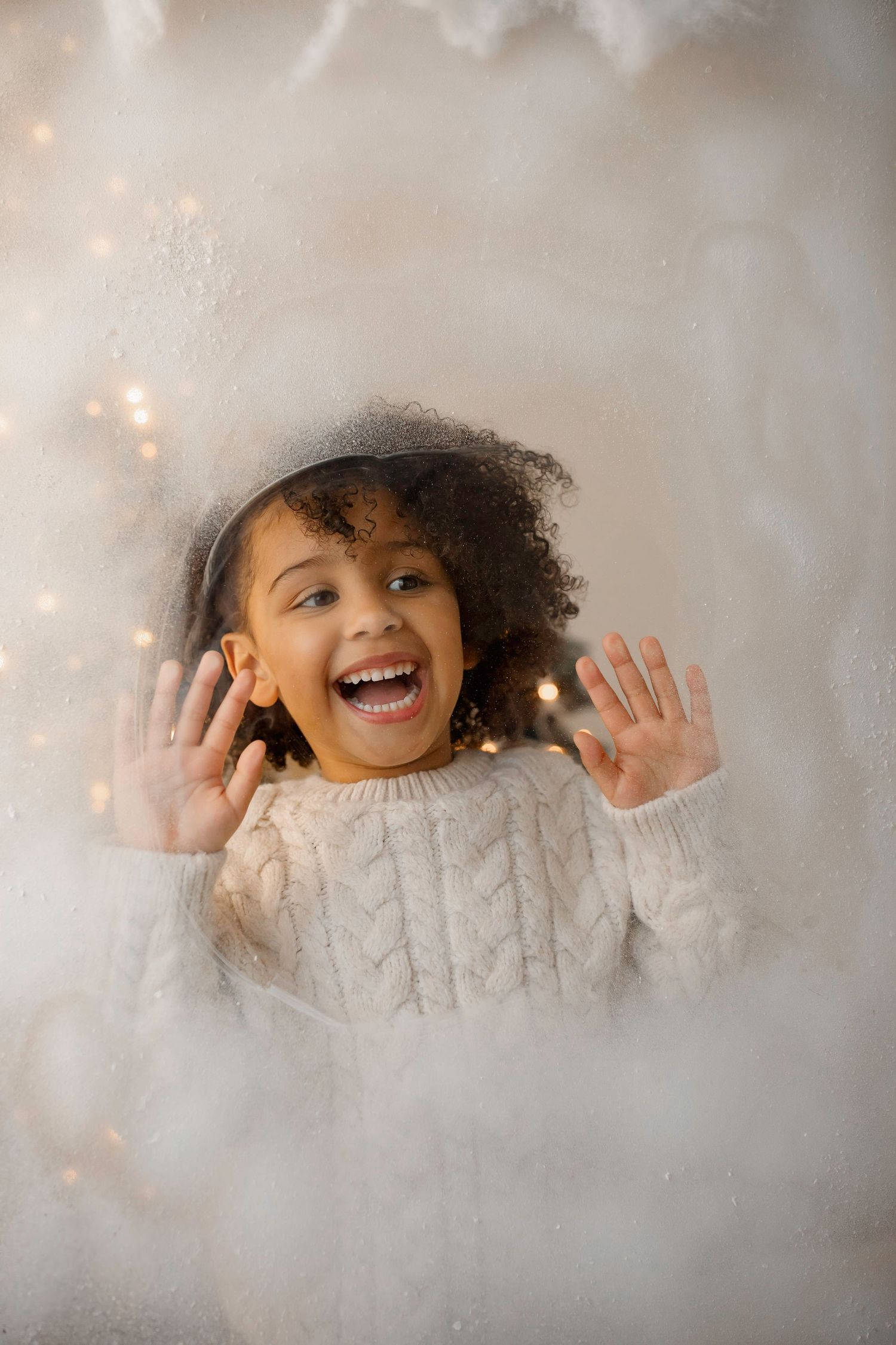 Young child in white sweater showing excitement with raised hands against a dreamy holiday background.