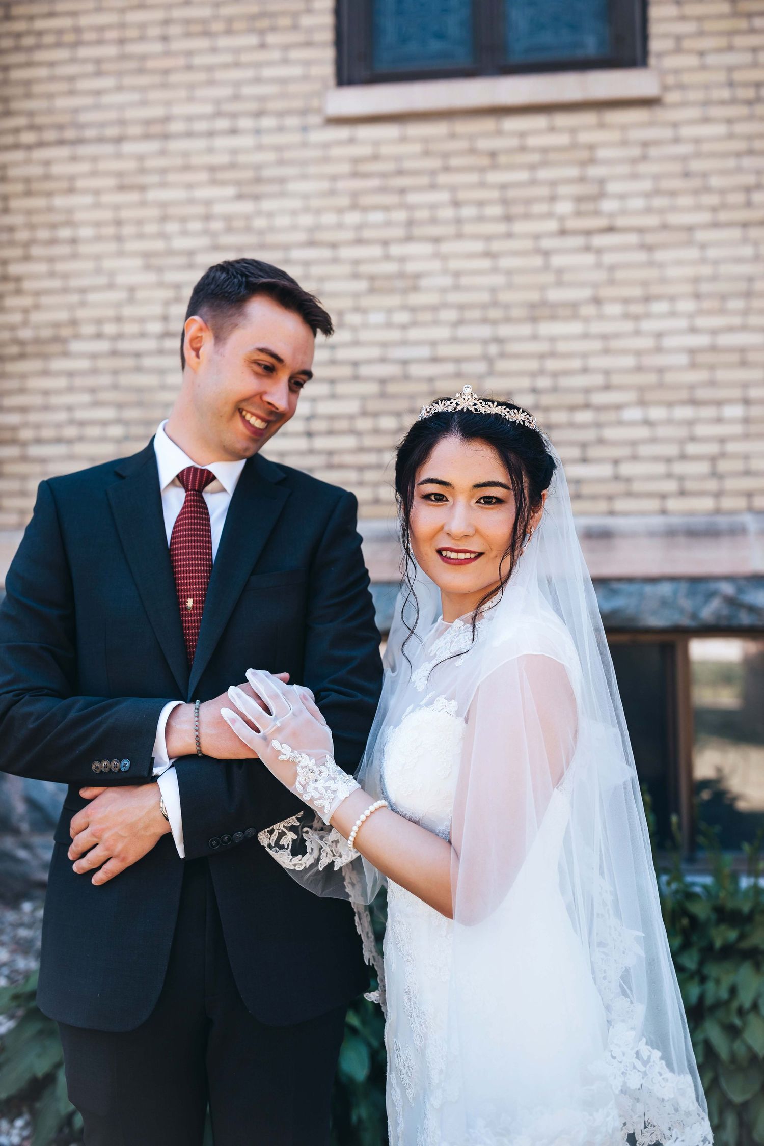 Elegant wedding portrait against brick church wall showing formal attire and veil.