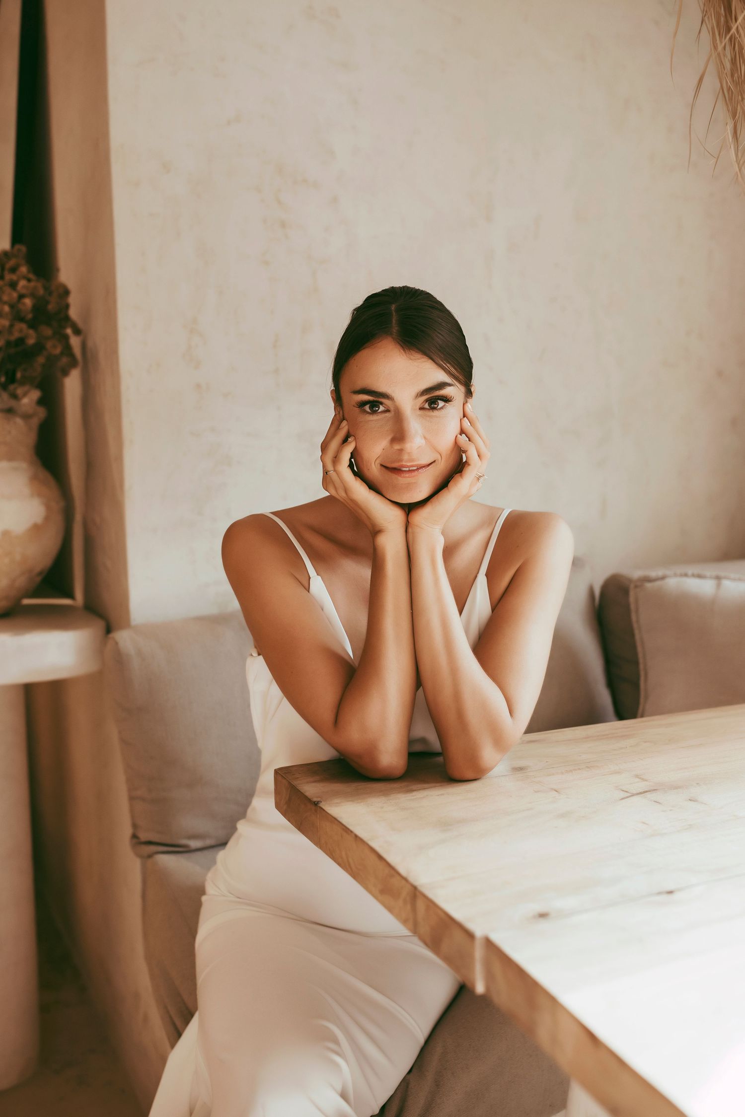 Woman in white dress resting chin on hands while seated at wooden table.